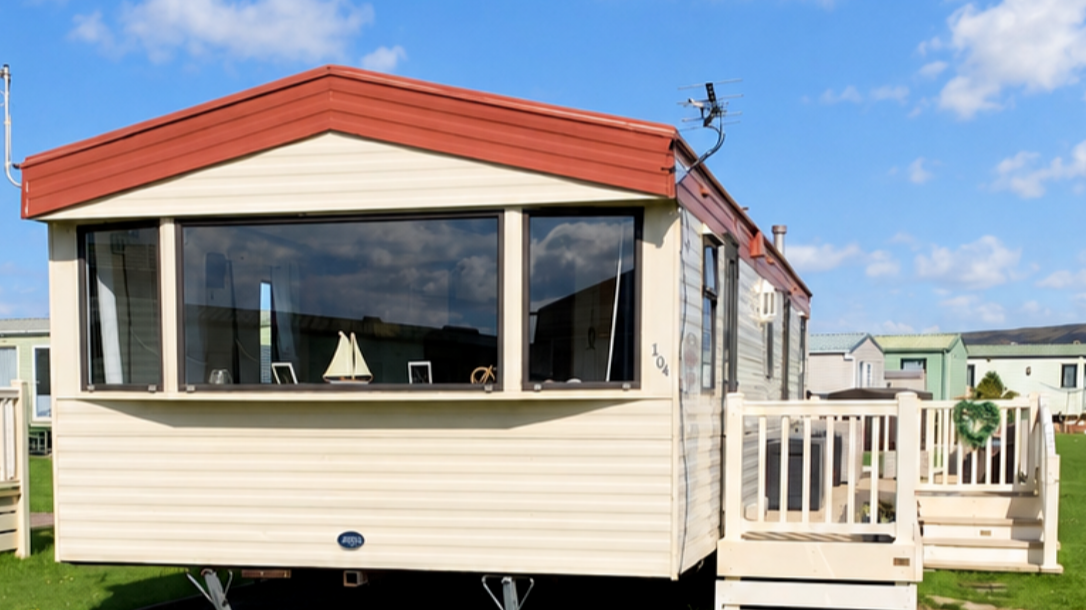 A beige mobile home with red roof in a caravan park, large windows showing a sailboat decoration and photos inside, small deck with white railing, green wreath decoration, surrounded by other mobile homes under a blue sky with clouds.