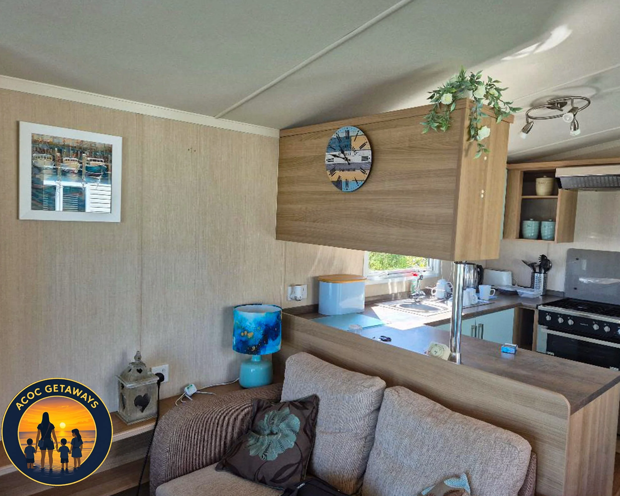 A cozy living room and kitchen area with beige seating, a blue lamp, a wooden dividing wall with a clock, a window, and kitchen appliances including an oven and shelves with dishes.