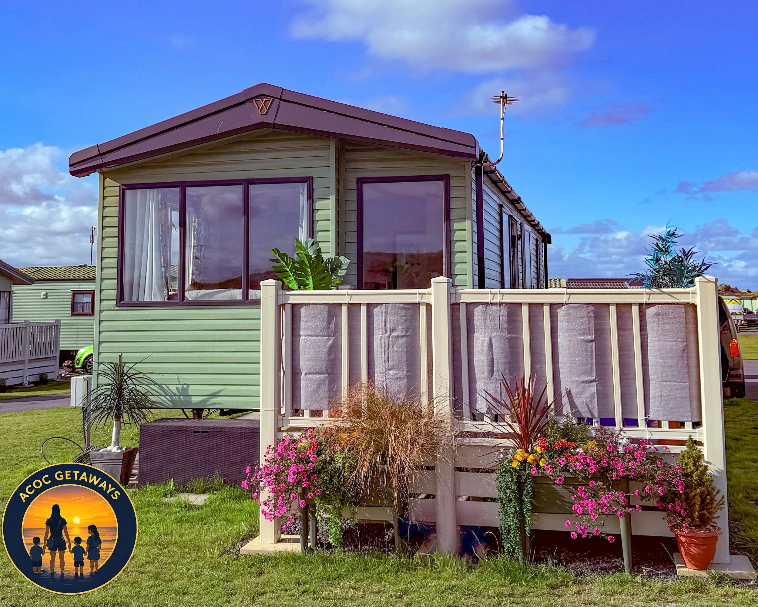 Cute mint green tiny house with brown roof, large front window with white curtains, and small enclosed porch with pink flowers and plants.