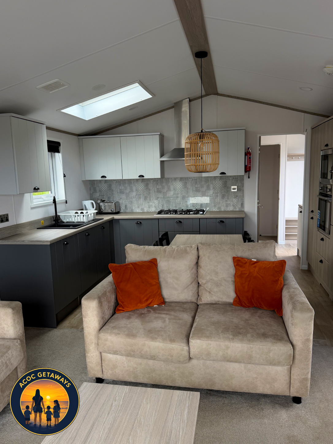 Open-concept living space with kitchen and beige sofa with orange throw pillows, skylight on ceiling, pendant light fixture, and doorway to another room.