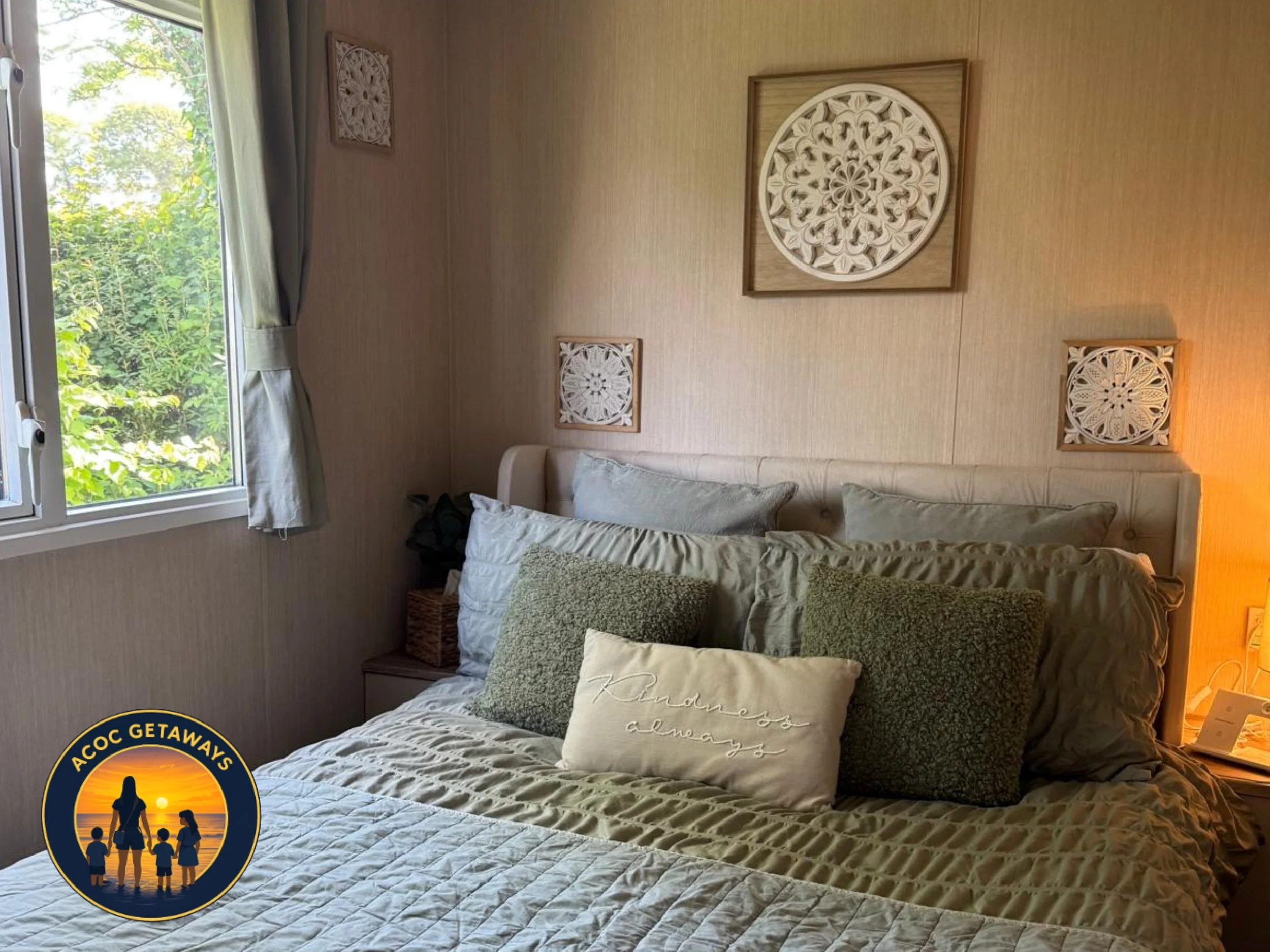 A bedroom with a bed covered in a beige quilt, multiple pillows including one with the text "Sometimes always," beside a nightstand with a lamp, and a window with light-colored curtains. Wall decor includes three intricately carved wooden panels arra