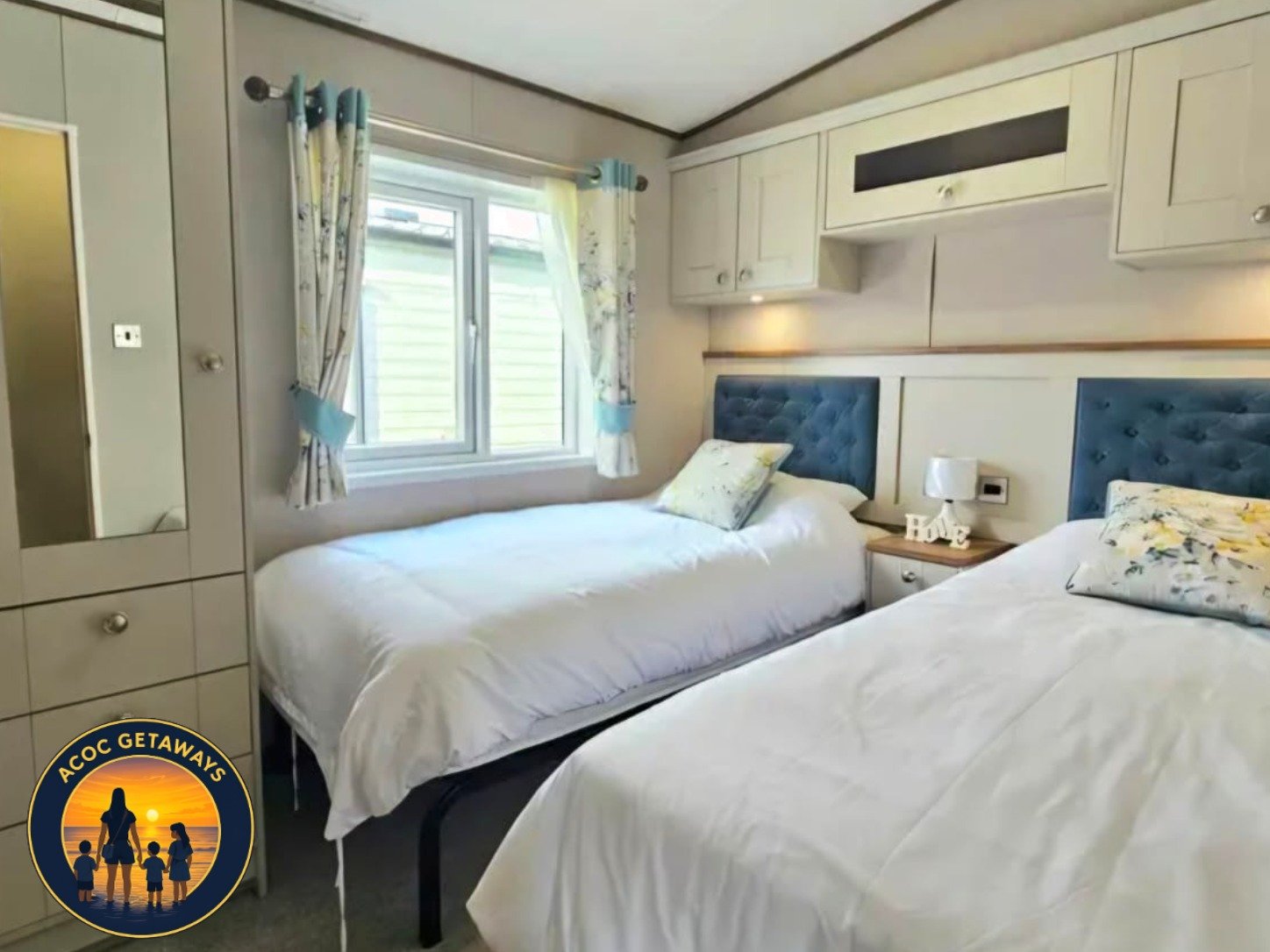 Two twin beds with white bedding and blue headboards in a cozy bedroom with a window, floral curtains, white cabinets, a bedside table with a small lamp, and decorative items.