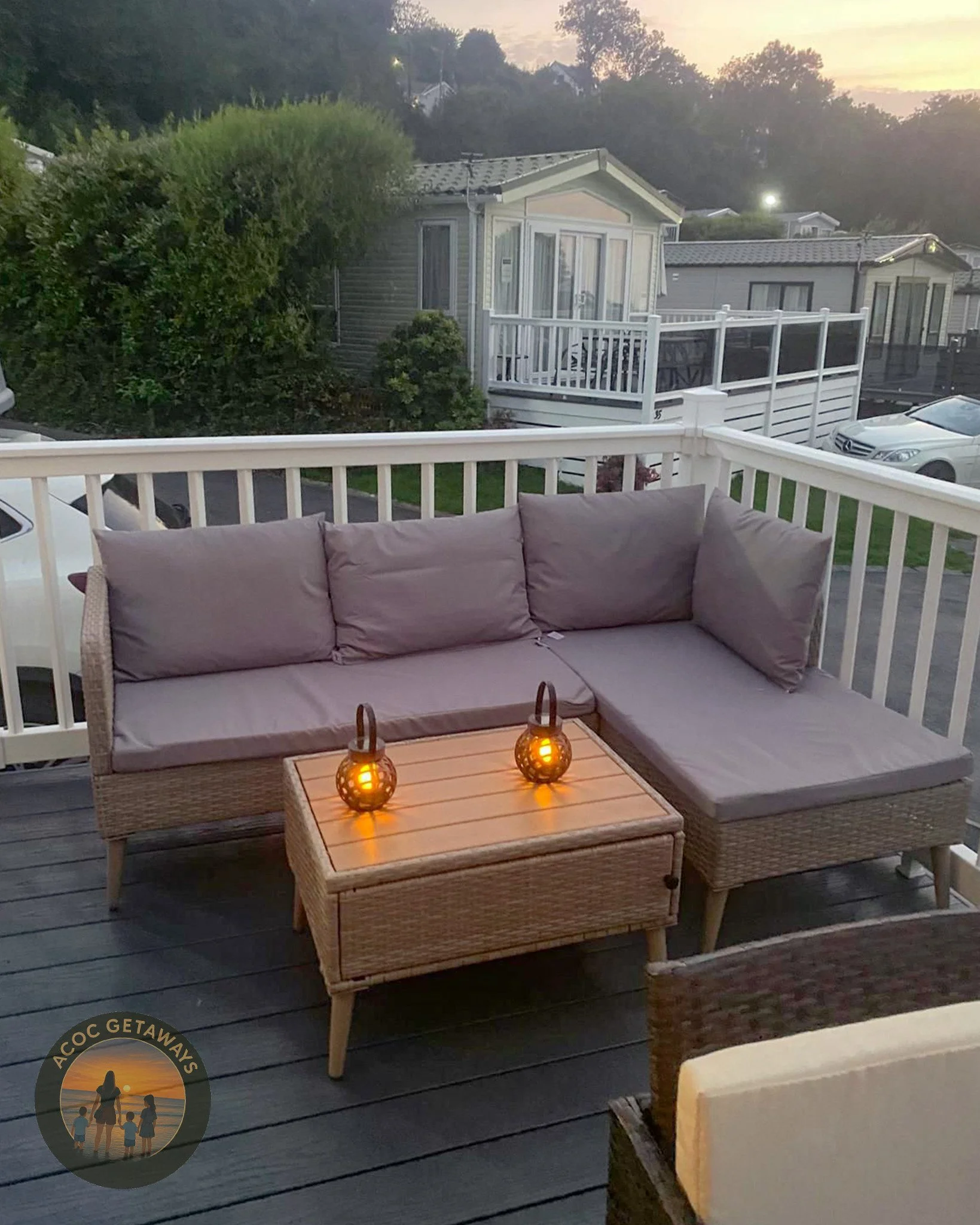 Outdoor deck with gray cushioned wicker sectional sofa, two glowing lanterns on a wooden table, surrounded by a white railing, overlooking neighboring mobile homes and trees at sunset.