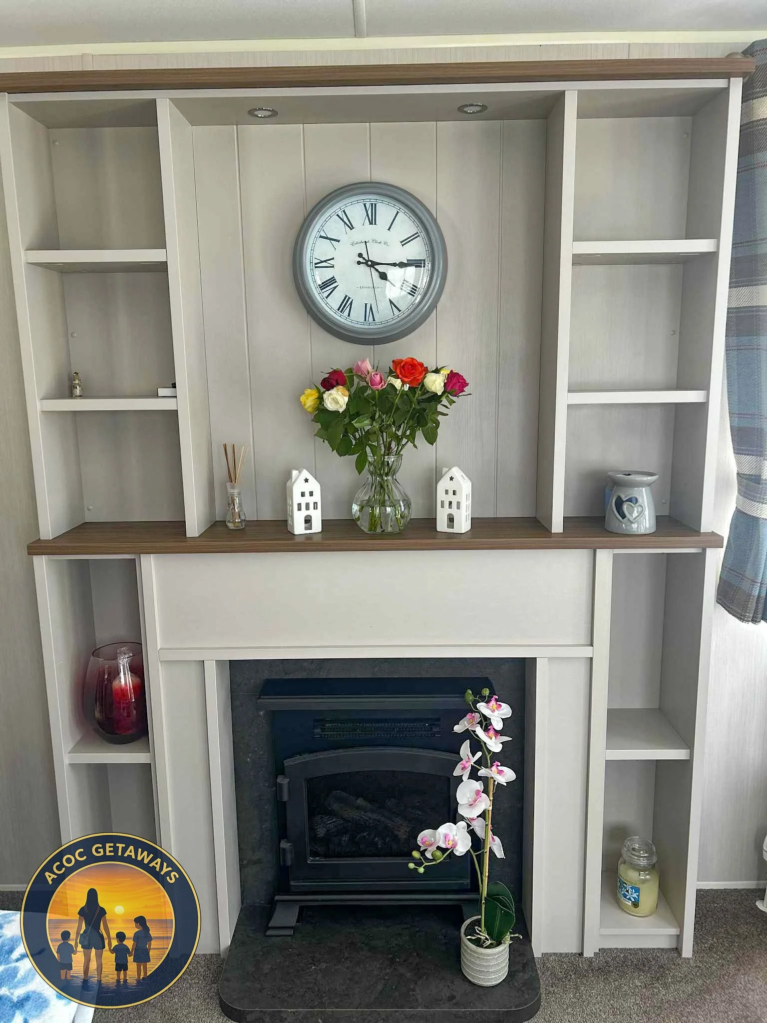 Decorative living room wall with a white wooden shelf, a round clock with Roman numerals, a glass vase of colorful roses, small candle holders, and various small decorative items. Fireplace with a black insert and a potted orchid in front.