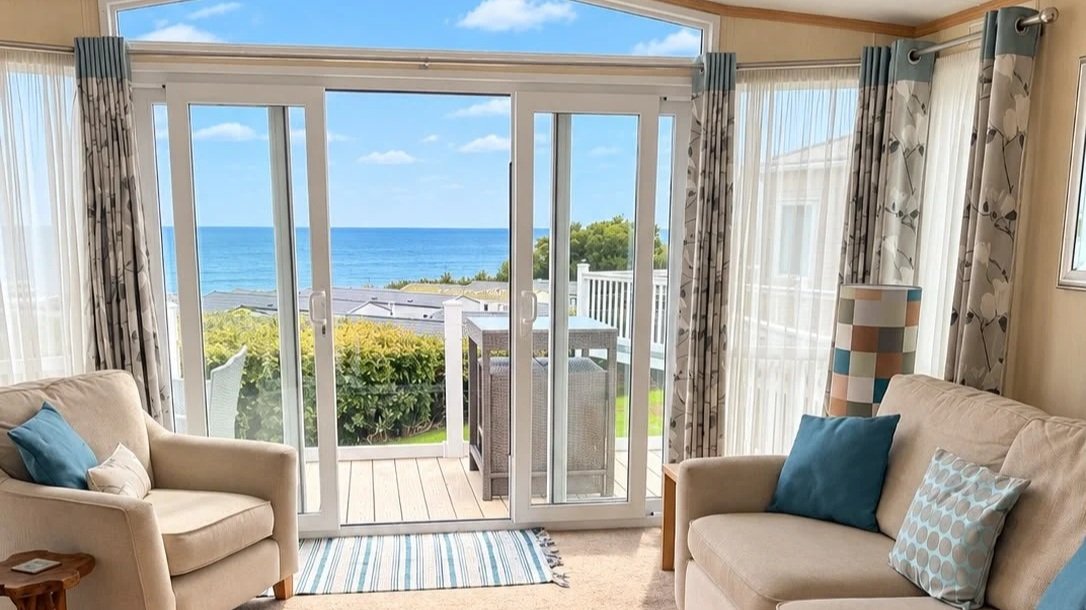 Living room with large sliding glass doors opening to a balcony, with a view of the ocean, blue sky, and greenery outside. Inside, there are two beige sofas with blue and patterned cushions, curtains with a leaf pattern, and a colorful lamp.