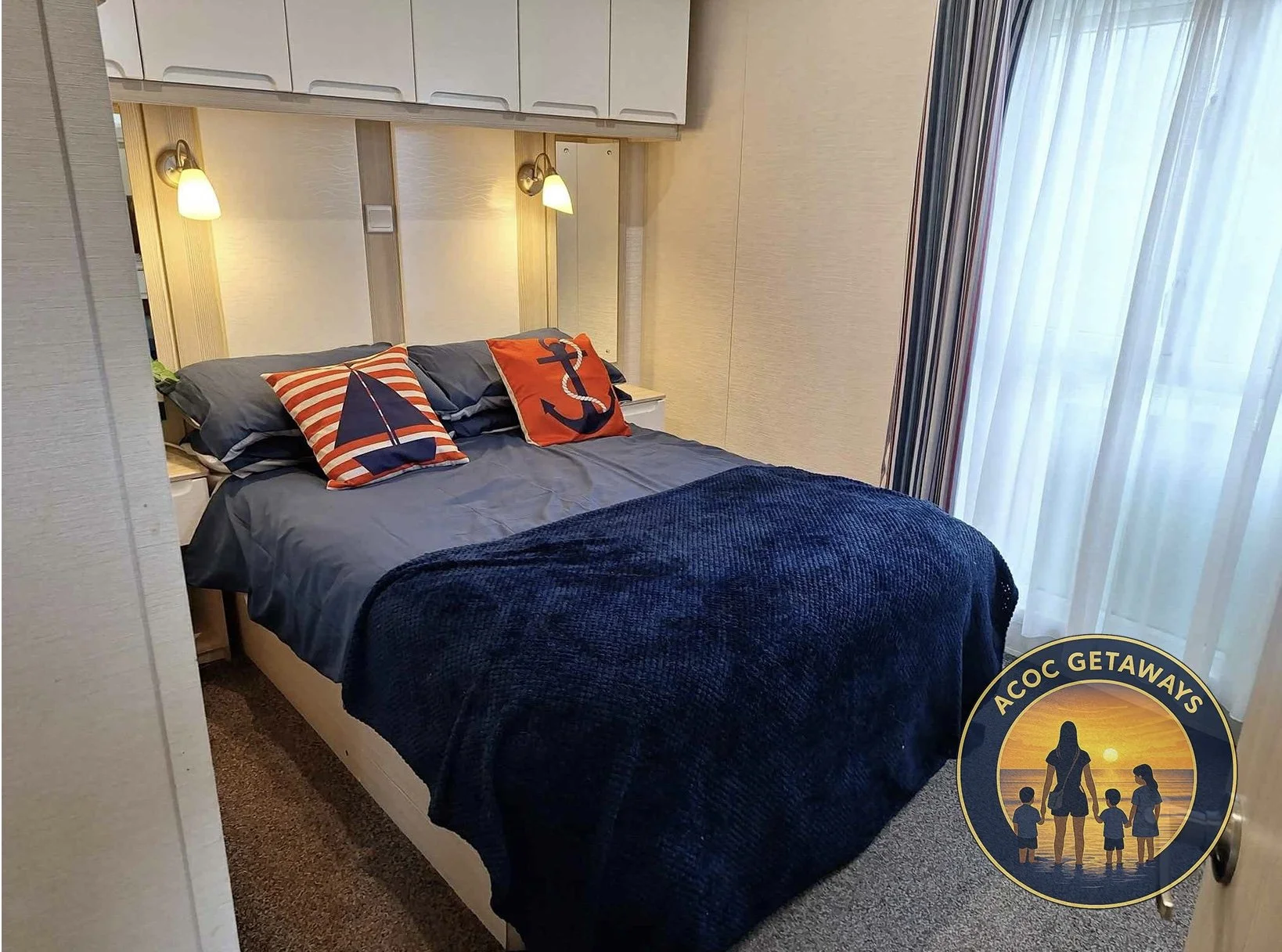 A neatly made bed with navy blue bedding and two decorative pillows with nautical designs, in a cozy hotel room with wall-mounted lighting and a large window with curtains.
