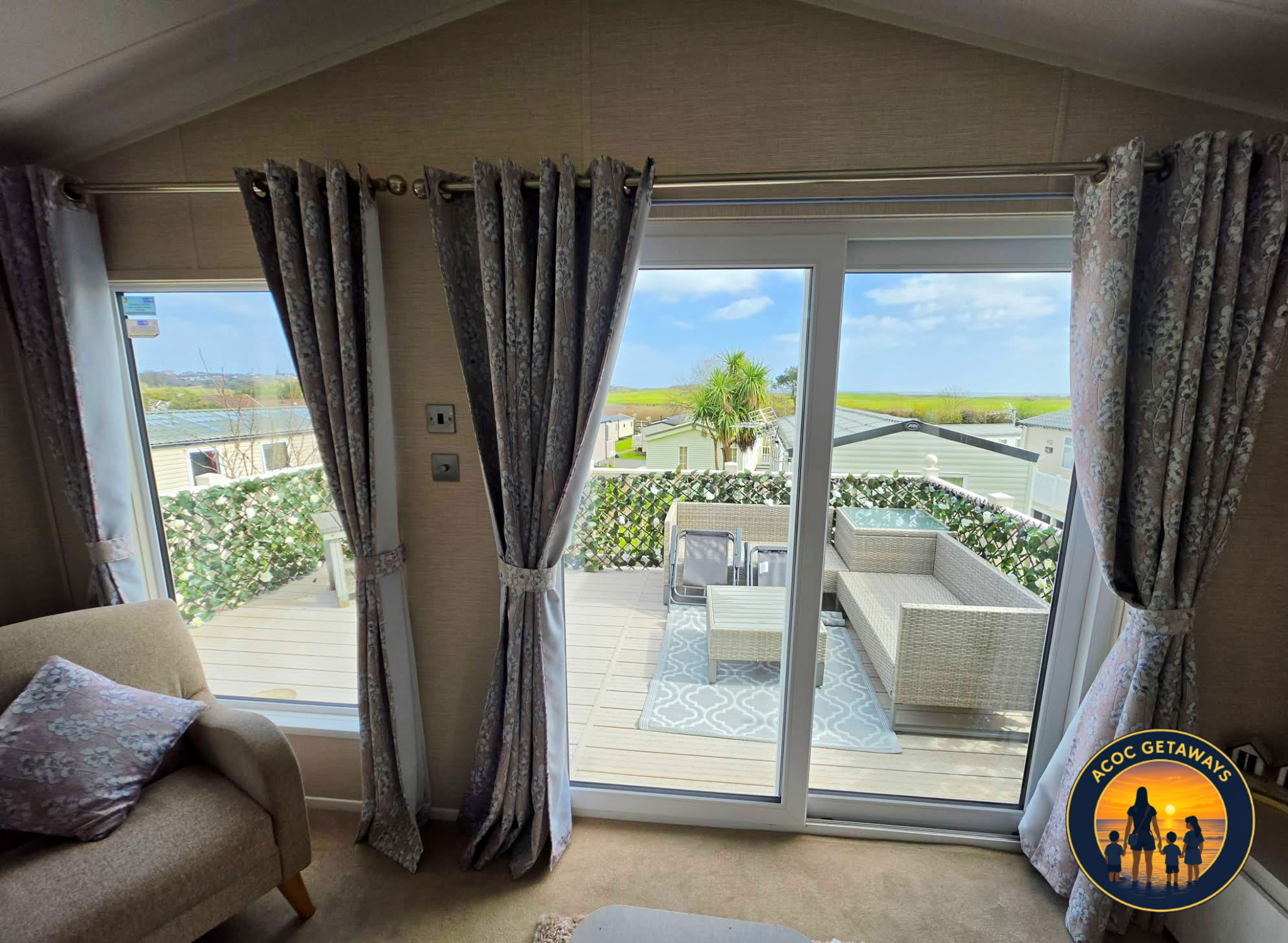 Living room with beige armchair, floral pillows, and sliding glass door leading to a patio with outdoor furniture. The patio overlooks a neighborhood with houses, greenery, and a bright blue sky with clouds.