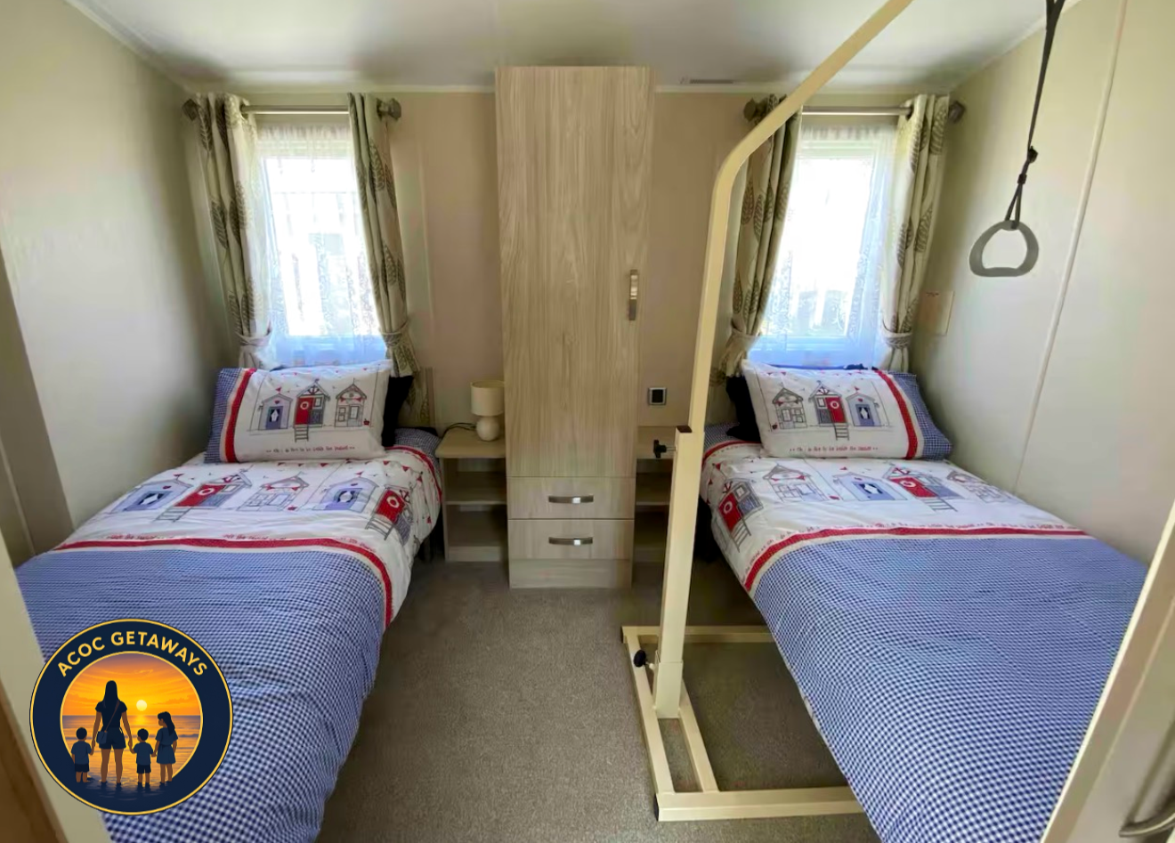A small bedroom with two twin beds, nautical-themed bedding, a window with patterned curtains, a nightstand with a lamp, and a privacy curtain divider.