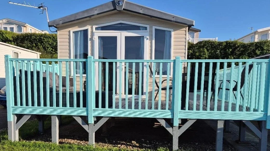 A small beige house with a wooden deck painted light blue, featuring a railing and outdoor chairs, against a clear blue sky.