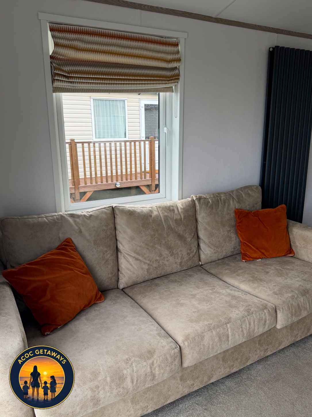Living room with a beige couch and orange pillows, window with a partial view of a wooden deck outside, dark blue vertical radiator on the right.