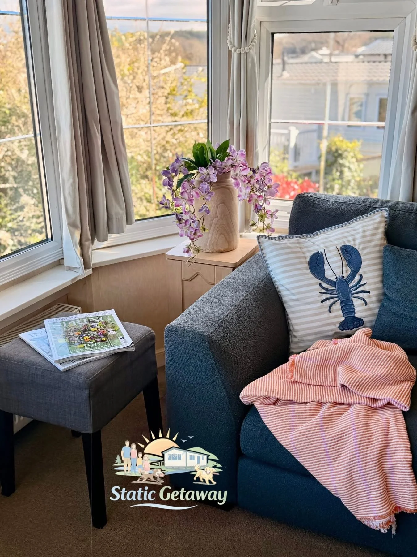 A cozy living room with large windows showing an outdoor view of trees and rooftops. On a small wooden side table, there is a vase with pink and purple orchids. A gray upholstered ottoman has a newspaper on it, and a couch with a plush pillow featuri
