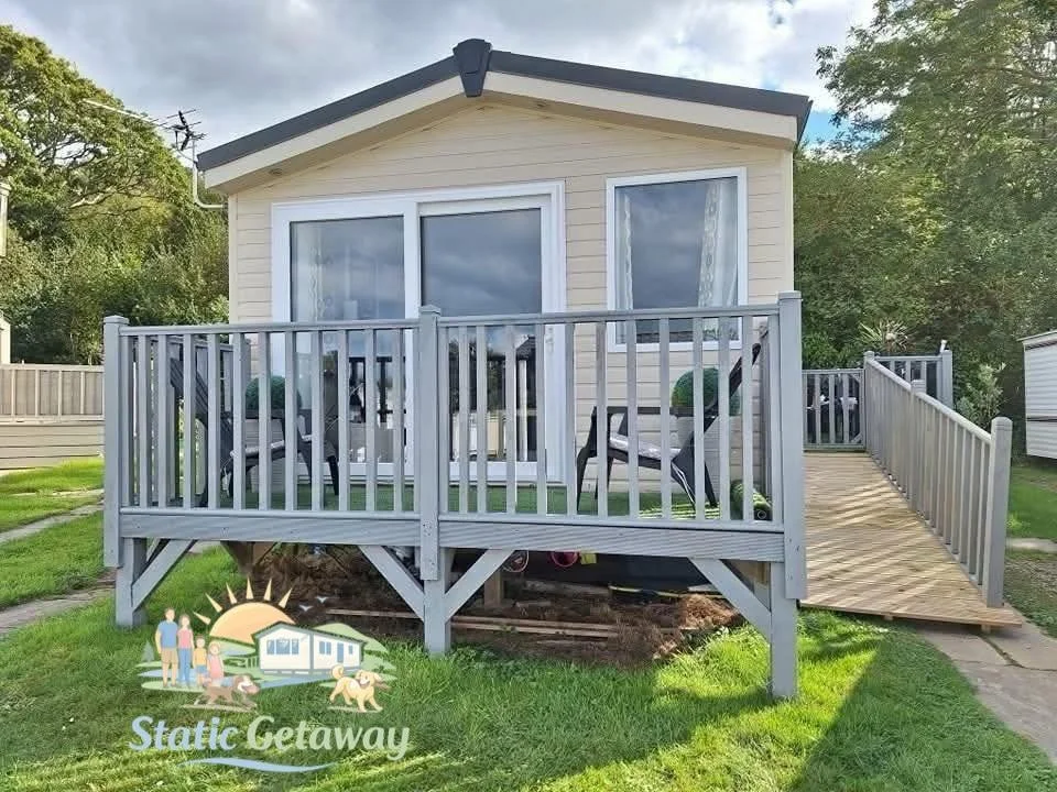 Small beige house with a wooden deck and white railing, surrounded by green grass and trees.