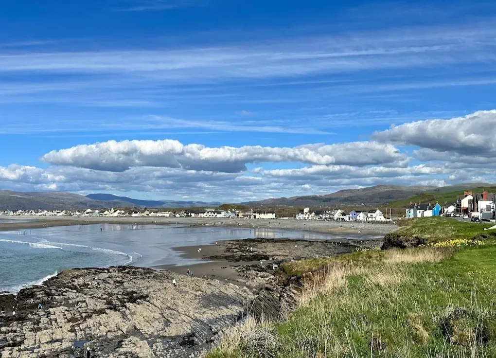 Coastal scene with rocky cliffs, sandy beach, and calm water. Small village with colorful houses along the shoreline under a partly cloudy sky.