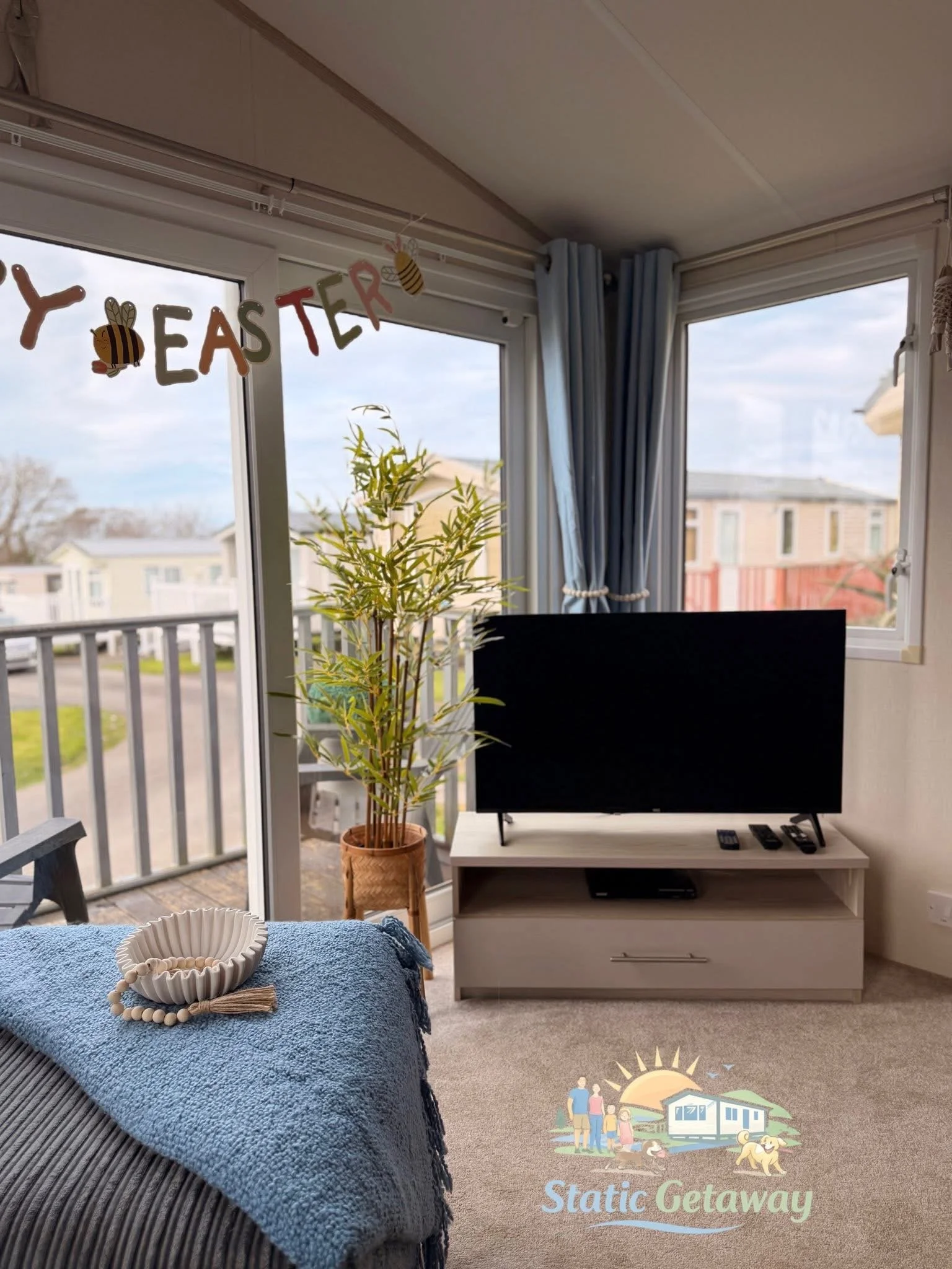 View from inside a living room or porch area with a sliding glass door leading to an outdoor balcony with railing. Inside, there is a television on a white stand, blue curtains, a potted bamboo plant, and a table with a blue textured blanket and deco