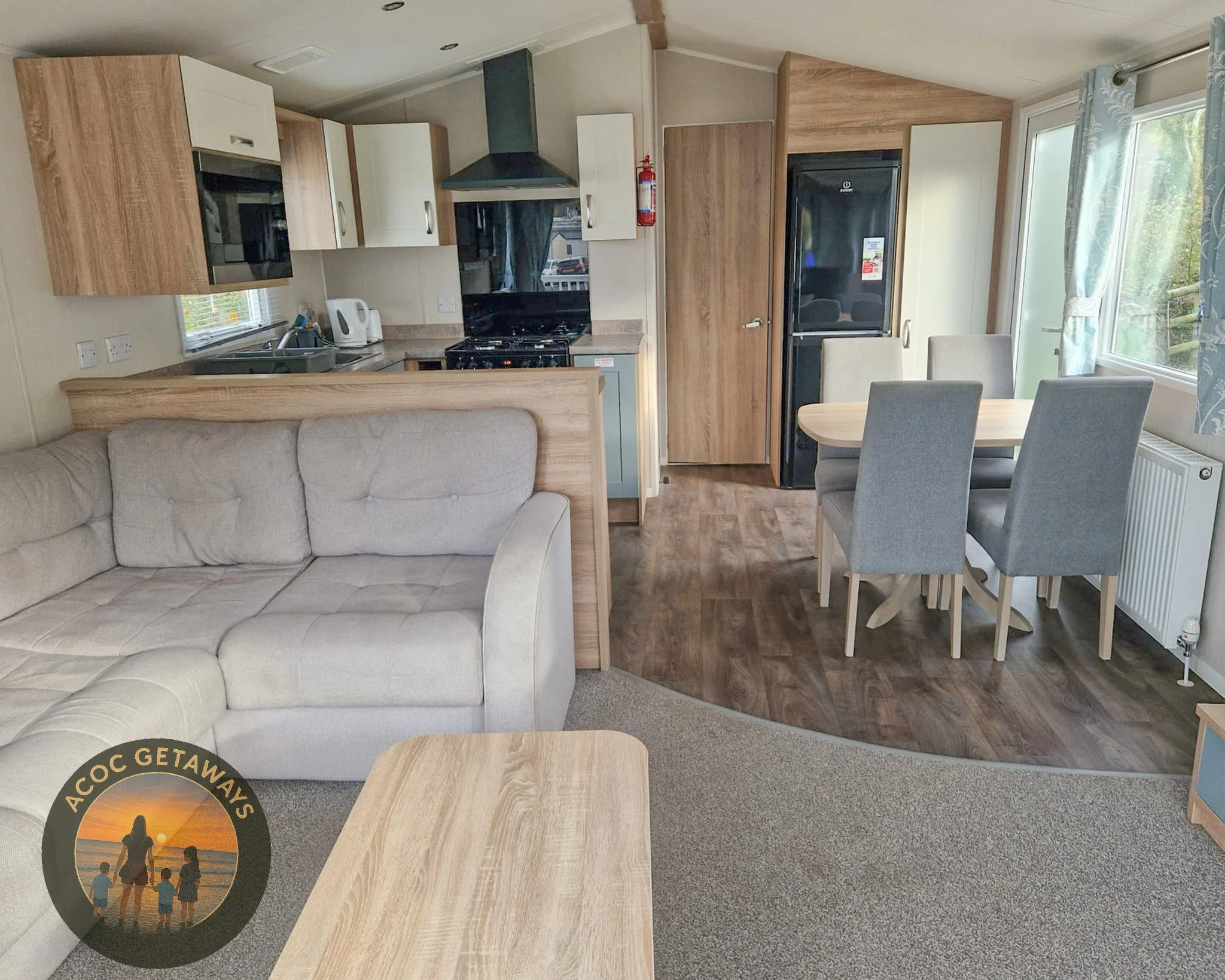 Interior of a cozy living room and kitchen in a holiday cabin, with a beige sofa, wooden dining table with gray chairs, compact kitchen area with black stove, refrigerator, microwave, and kettle, and large windows with blue patterned curtains showing a view of greenery outside.