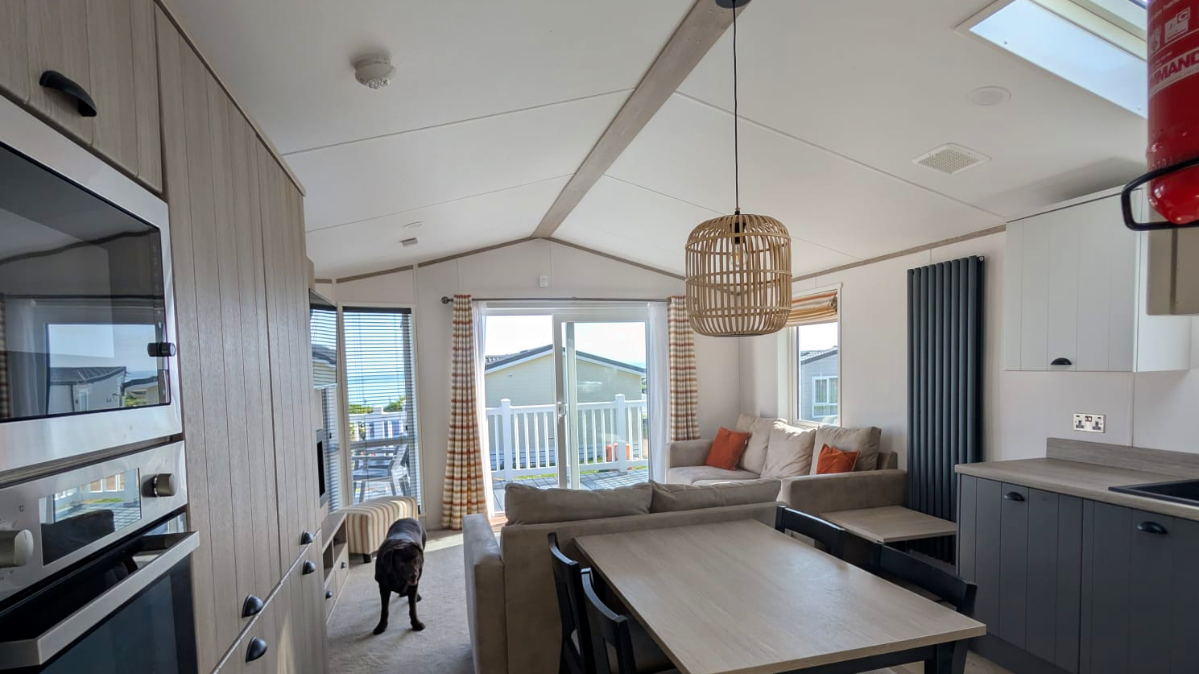Interior of a cozy living room with a beige sectional sofa, orange and white throw pillows, a wooden dining table with black chairs, and a kitchen area. There are sliding glass doors leading to a balcony with outdoor furniture, and a black dog is standing in the room. The space is bright with natural light and has light-colored walls and ceiling.