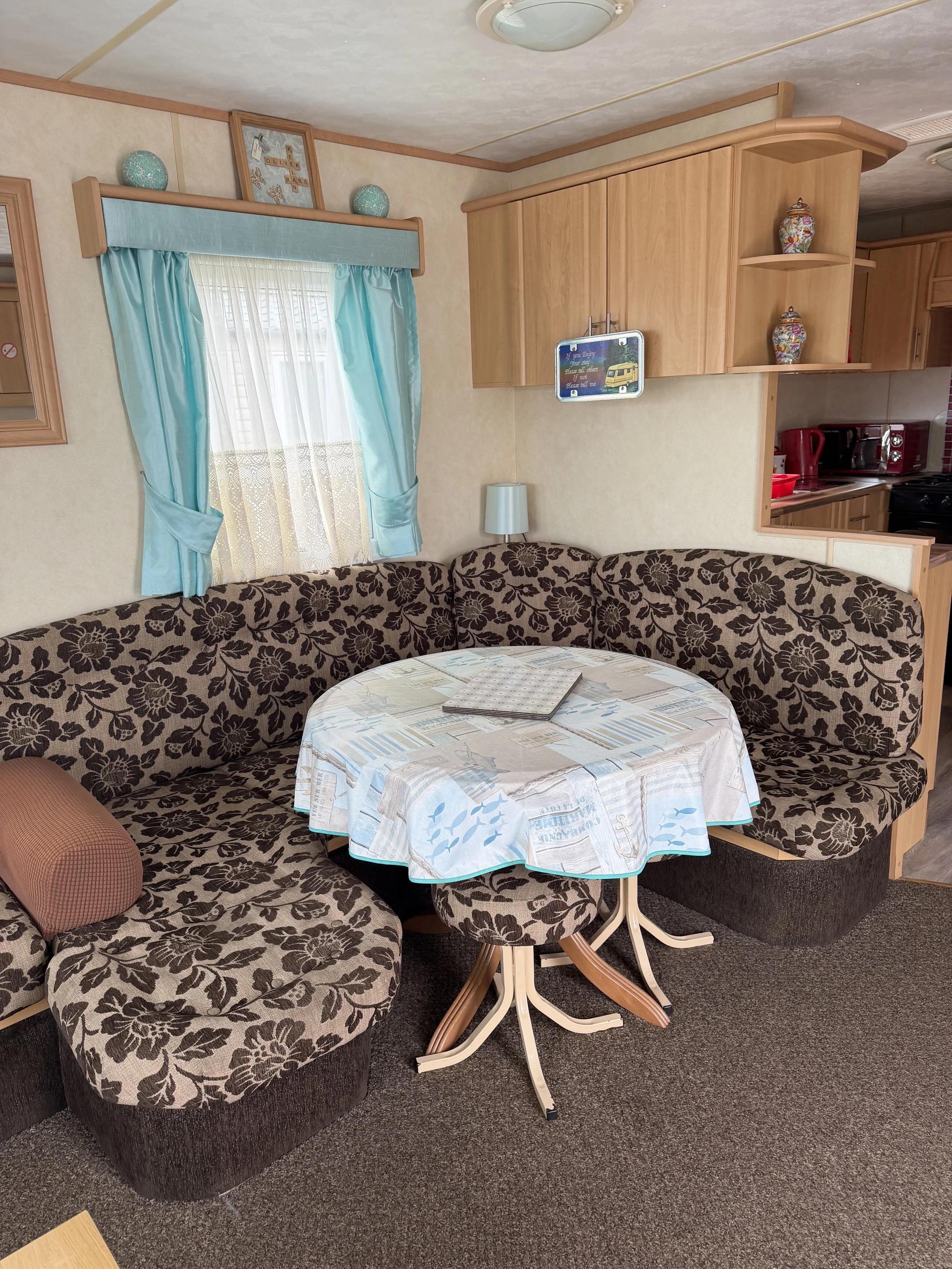 Living room with brown floral patterned sectional sofa, small round dining table with a nautical themed tablecloth, light blue curtains over window, wooden cabinets, decorative vases on shelves, and a small lamp in the corner.