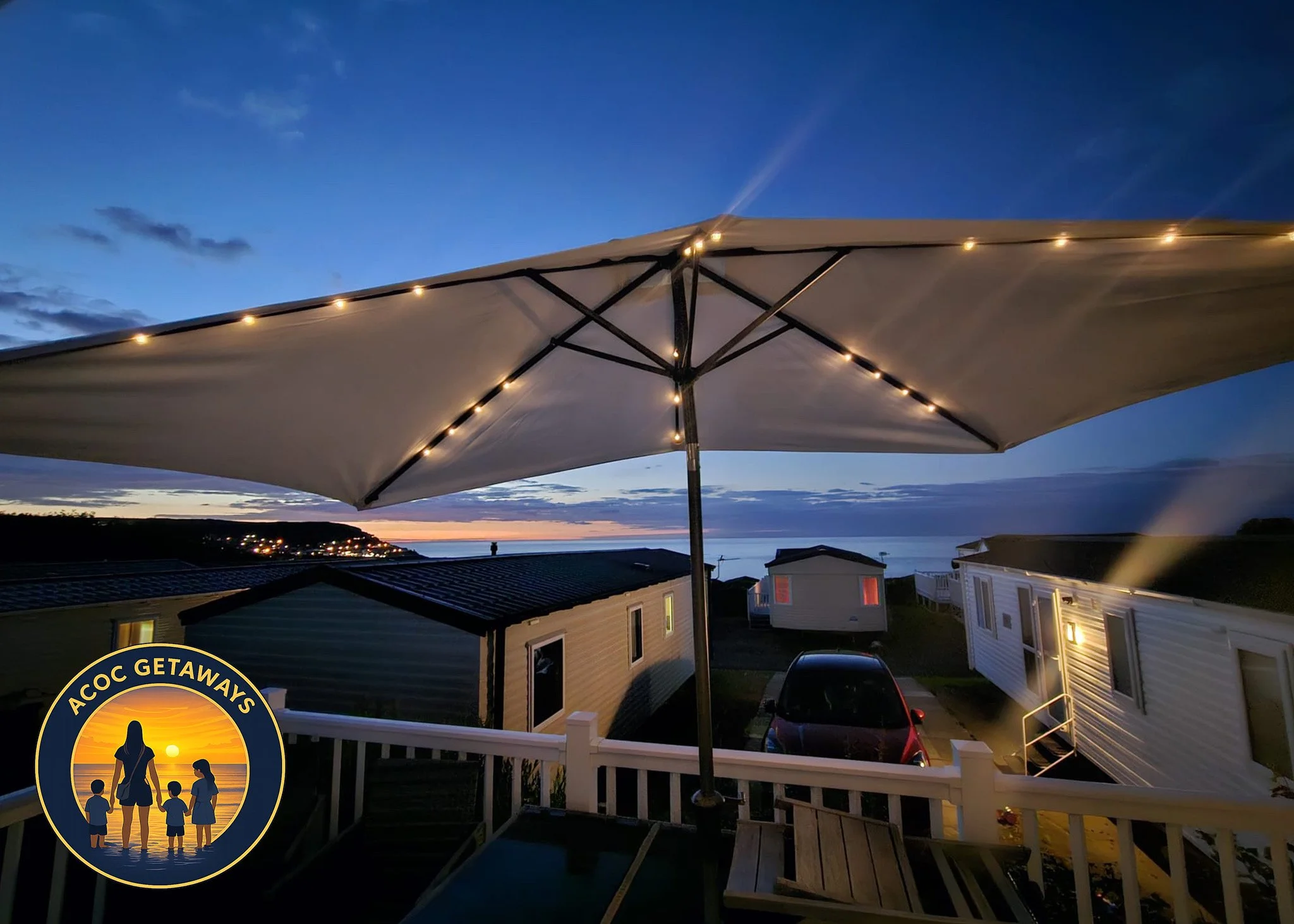 A view of a seaside campsite at dusk with an illuminated large outdoor umbrella on a deck, surrounding small mobile homes, and a car parked nearby, overlooking the ocean and a distant illuminated shoreline.