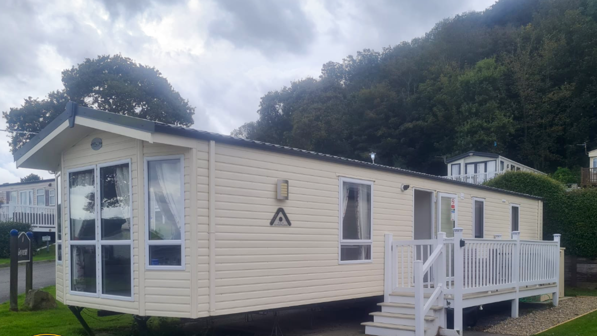 A beige prefabricated mobile home with white trim, a small porch with white railing, and several windows, situated in a park area with trees and other similar homes in the background.