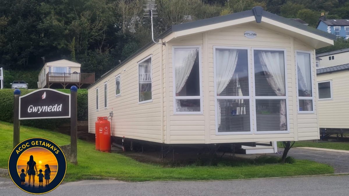 A beige mobile home with white curtains, set on a grassy area near a road, with a sign that reads "Gwynedd" and an ACOC Getaways logo in the corner.
