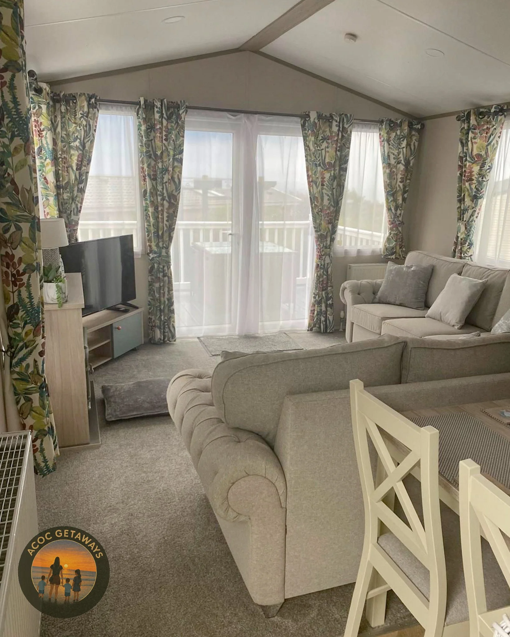 Living room with beige sofas, floral curtains, a TV on a wooden stand, and large glass door with sheer and floral curtains leading to a balcony.