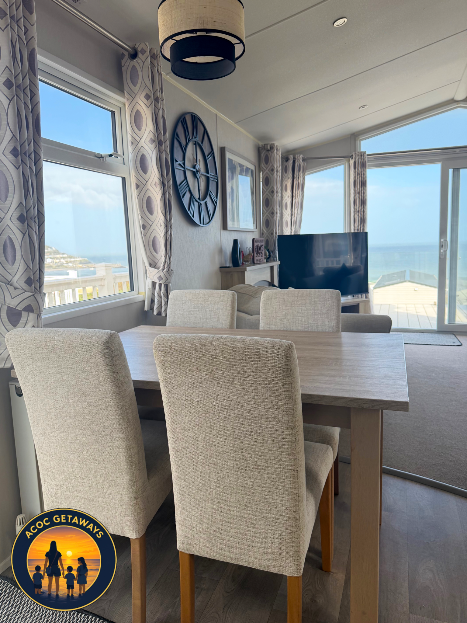 A dining area with beige upholstered chairs around a light wooden table, large windows with patterned curtains, a wall clock, and a flat-screen TV, with ocean views in the background.