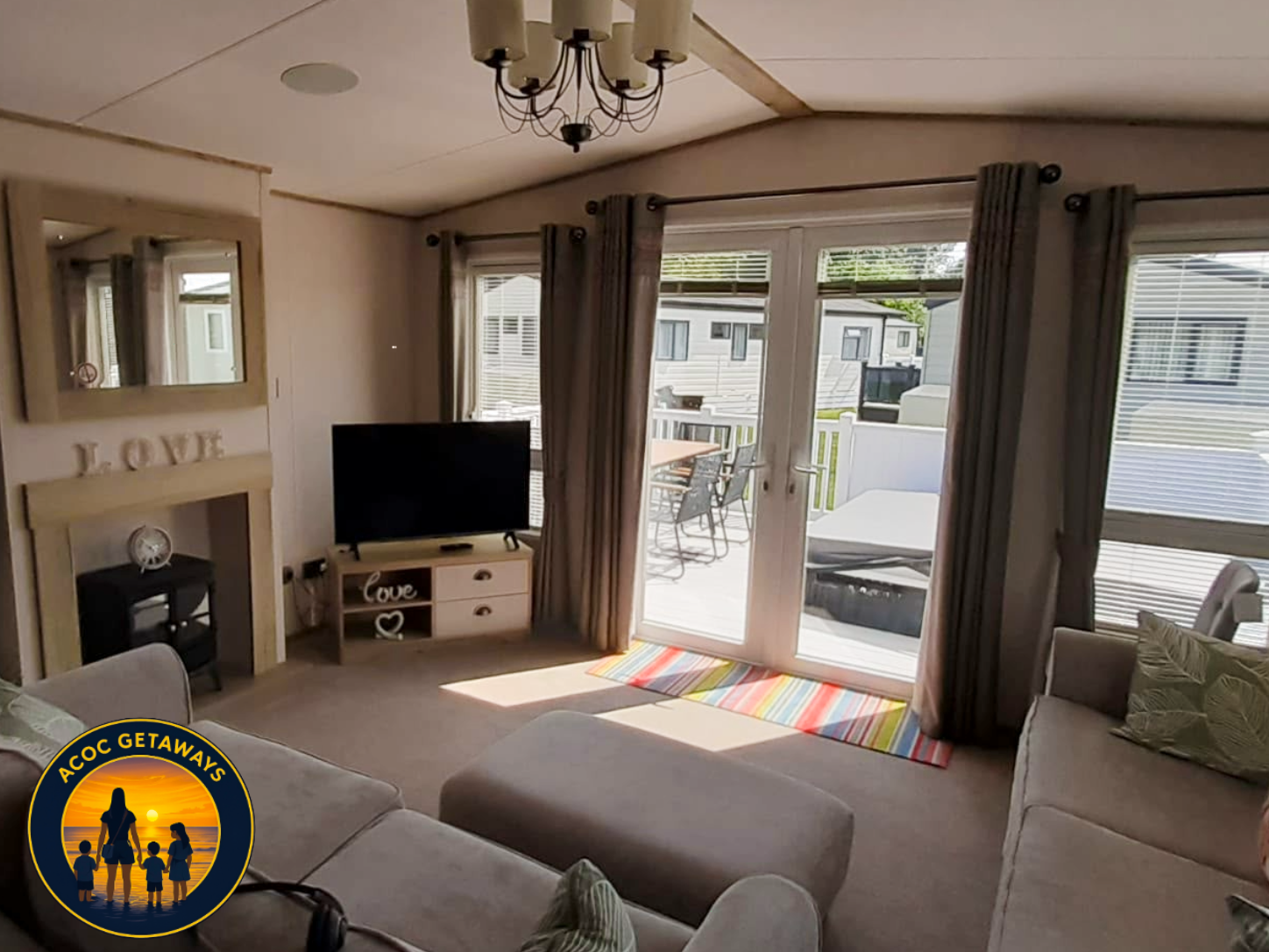 Living room with a large beige sectional sofa, a black flat-screen TV on a small white cabinet, a colorful striped rug, and sliding glass doors that open to a patio with outdoor furniture and neighboring trailers.