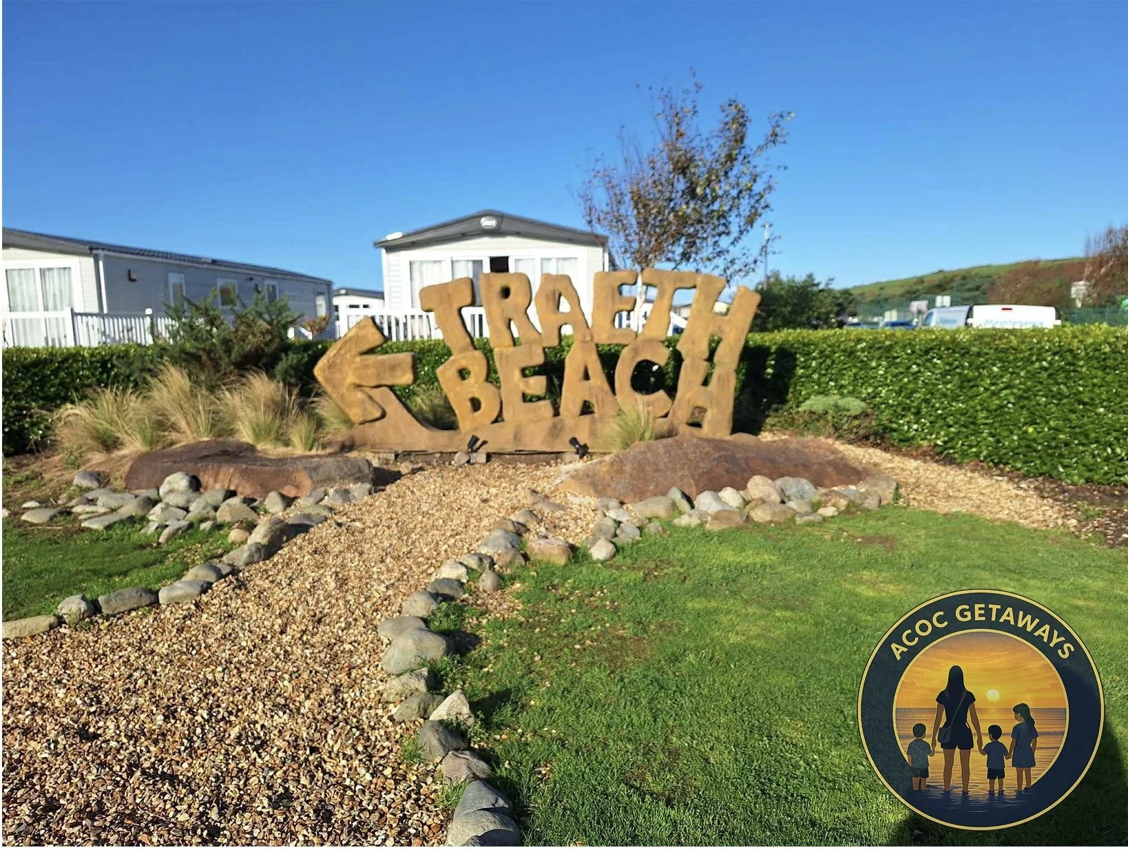 Sign at Trafalgar Beach with large stone letters and an arrow pointing left, against a backdrop of mobile homes, a tree, and a hedge, with a blue sky; inset logo with silhouetted woman and children at sunset, reading "ACOC GETAWAYS."