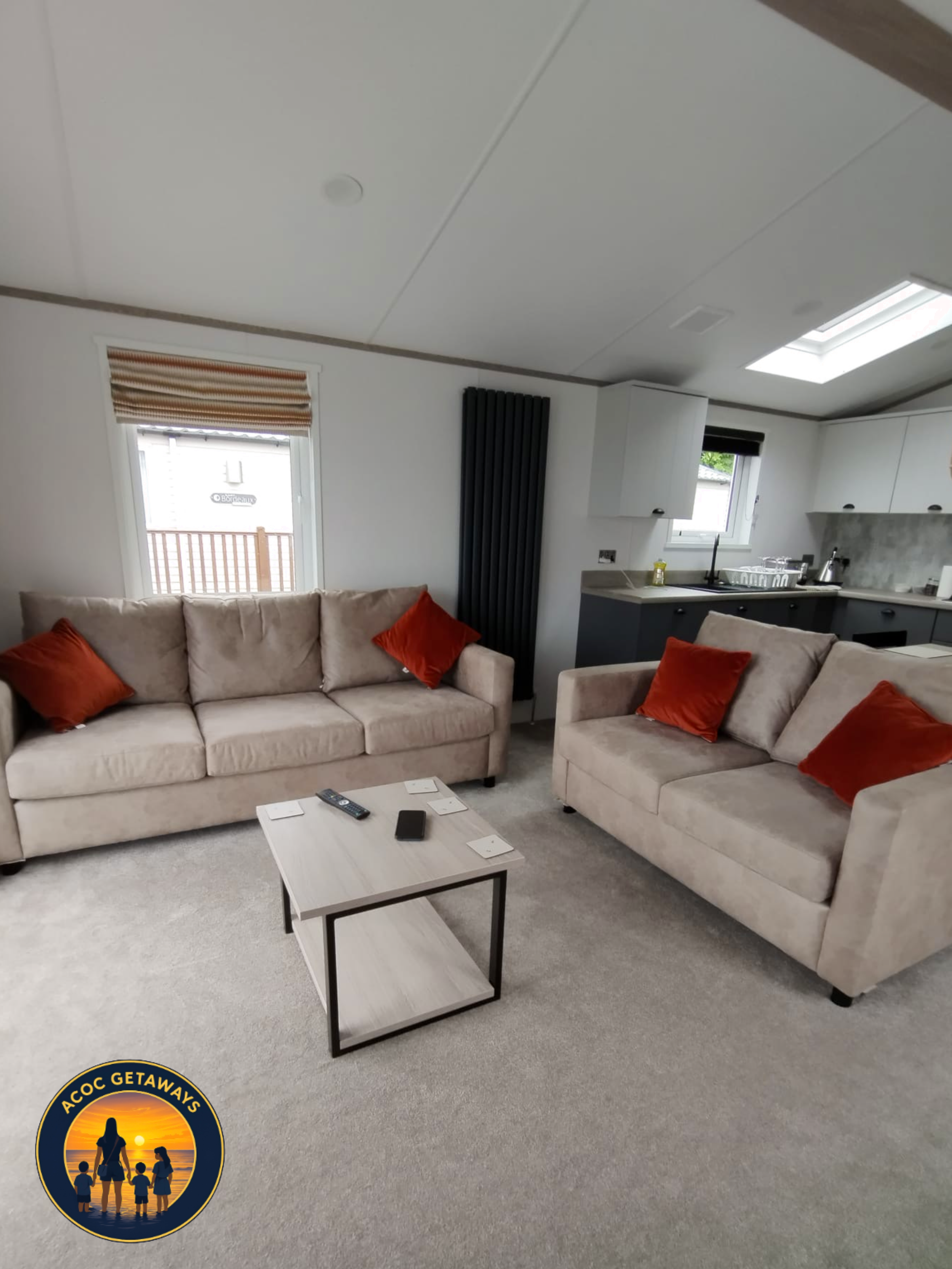 Living room with beige sofa and orange pillows, a small rectangular coffee table with remote control and coasters, gray plush carpet, kitchen area with white cabinets, black countertops, and black appliances, two windows, one with a brown-striped rom