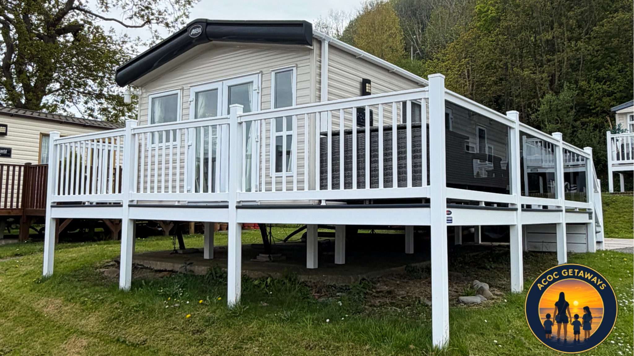 Backyard view of a house with a large white wooden deck and white fencing, with a hillside covered in trees in the background.