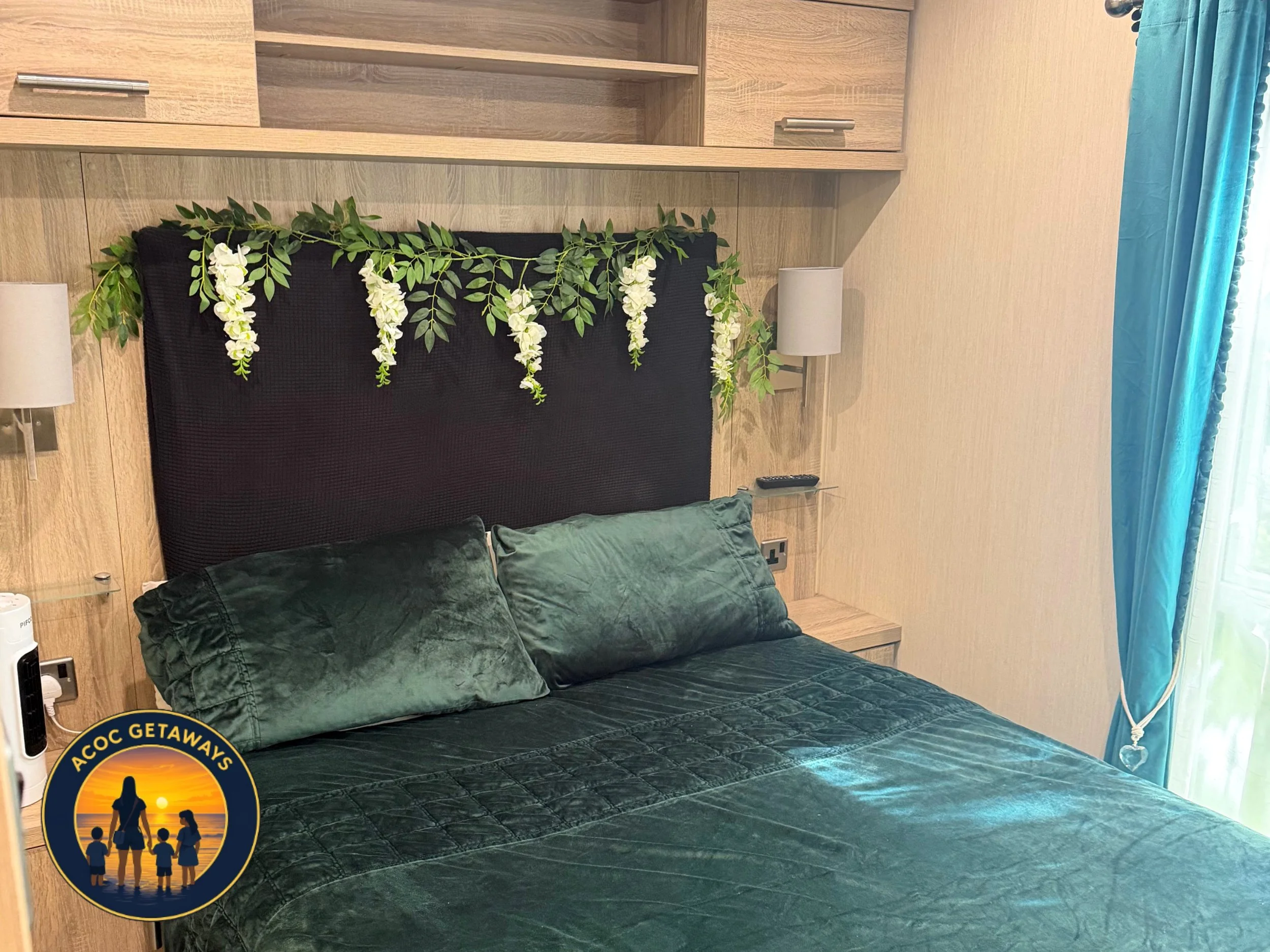 A bedroom with wood-paneled walls, a black headboard decorated with hanging white flowers and green leaves, and two green pillows on a dark green velvet bedspread. A white bedside lamp is on the right side of the bed, and a small corner shelf is buil