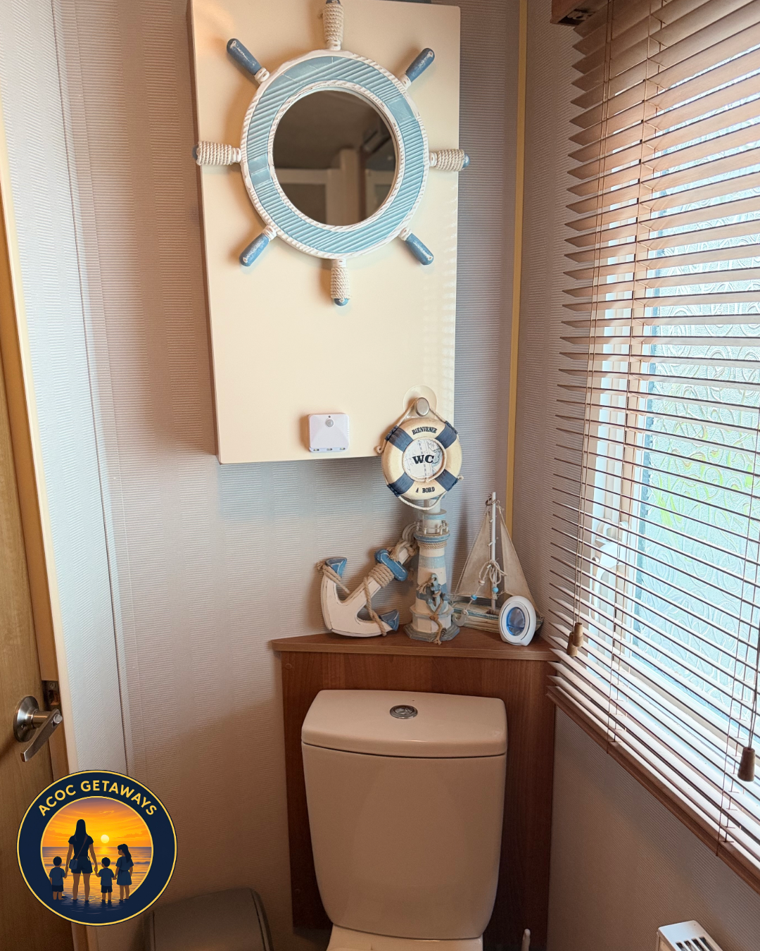 A small bathroom with a nautical themed decor, including a mirror framed like a ship's wheel, a life preserver-shaped sign, and boat and anchor decorations on the wall above the toilet, next to a window with blinds.
