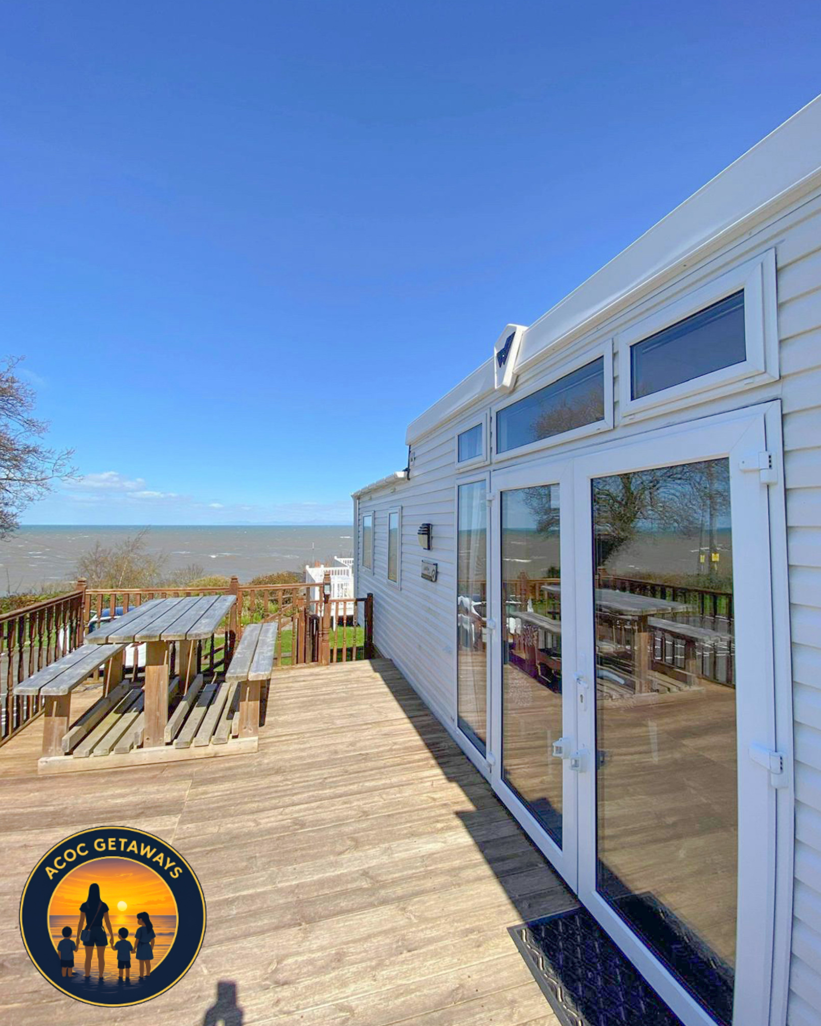 Beach house deck with wooden table and bench, railing, and large glass windows reflecting trees and sky, overlooking a sandy beach with the ocean in the background under a clear blue sky.