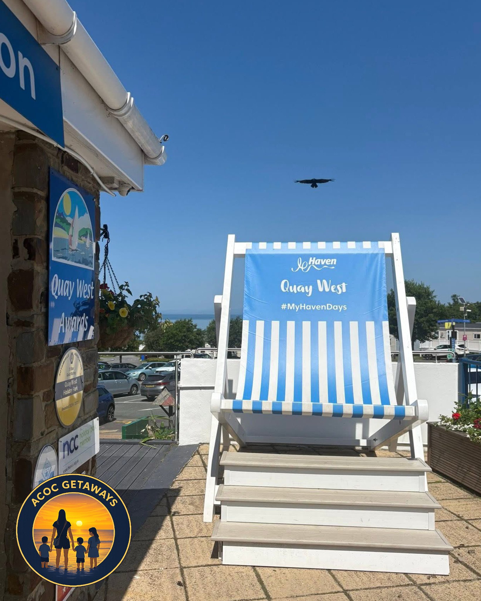 A sign on the left side reads "Quay West Awards" and features a photo of a sailboat. A large blue and white striped beach chair with a sign on top reads "Haven Quay West #MyHavenDays". In the background, a plane is flying in a clear blue sky. There a