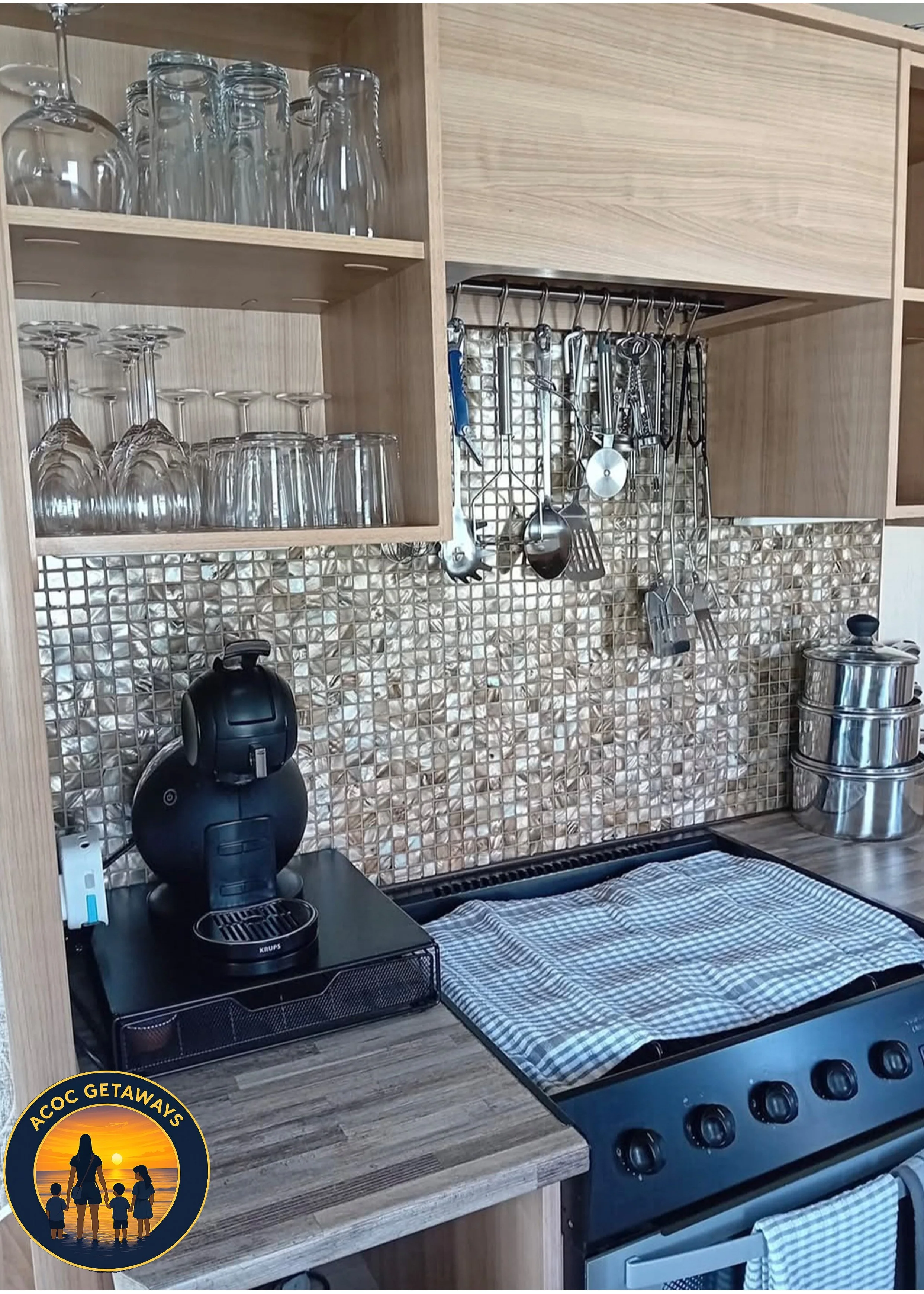 Kitchen corner with wooden shelves holding glasses, a black coffee machine on the counter, a stove with a checkered cloth, and a backsplash of small shiny square tiles.