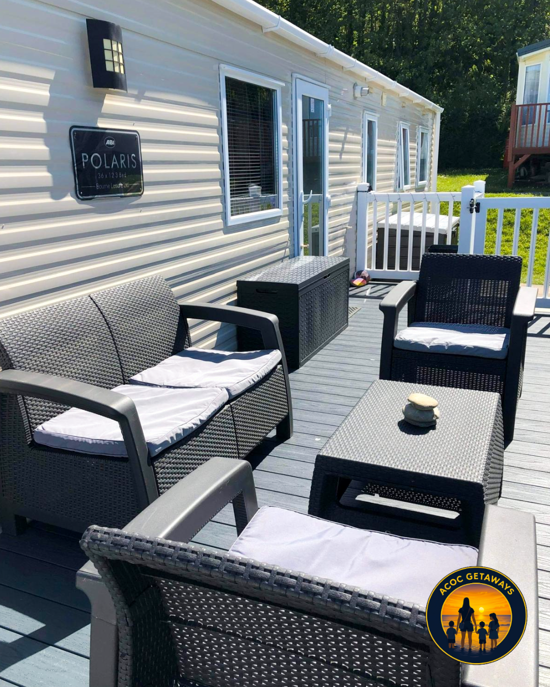View of a porch with outdoor furniture including a cushioned loveseat, two cushioned chairs, a small ottoman with stones on top, and a storage chest, all on a wooden deck adjacent to a mobile home with a sign labeled 'Polaris'.