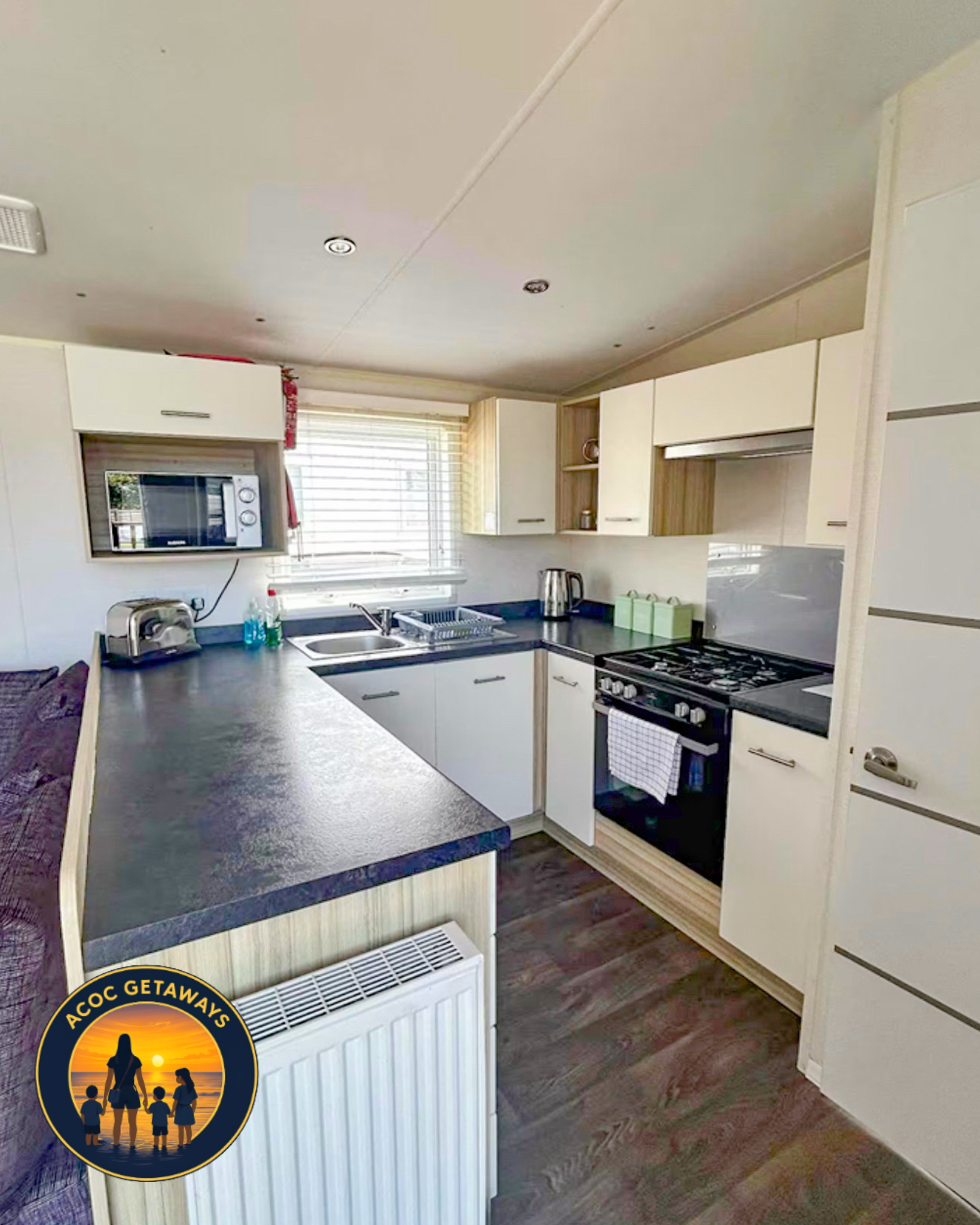A kitchen with white cabinets, a black countertop, a microwave oven, a dish rack, a kettle, a sink, window with blinds, and a wooden floor.
