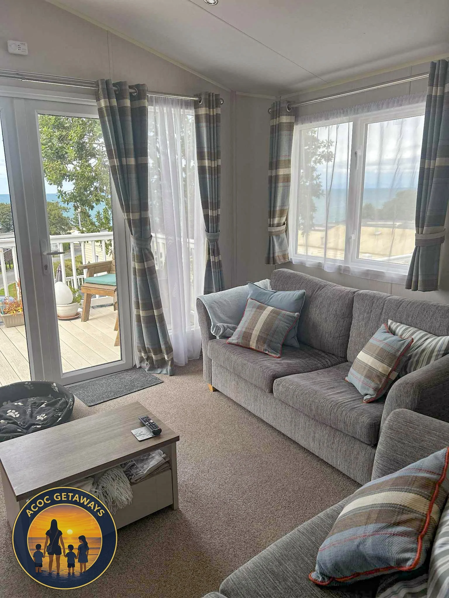 Living room with a gray sofa and plaid pillows, a coffee table with remote control, and large windows with curtains and a sliding glass door leading to a balcony with outdoor furniture, overlooking trees and water.