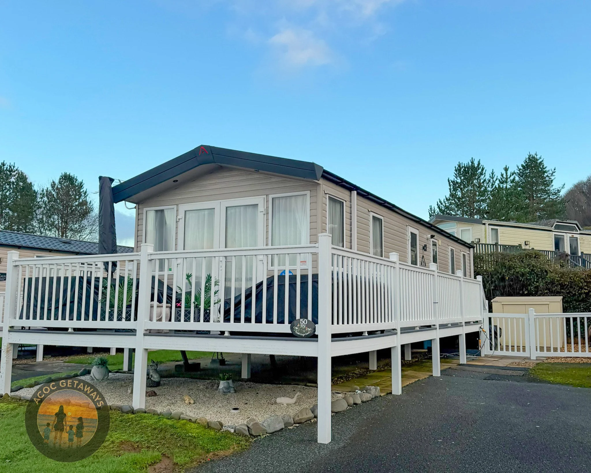 A beige mobile home with a white railing deck, sliding glass door, and several windows, located in a mobile home park with trees in the background during daytime.