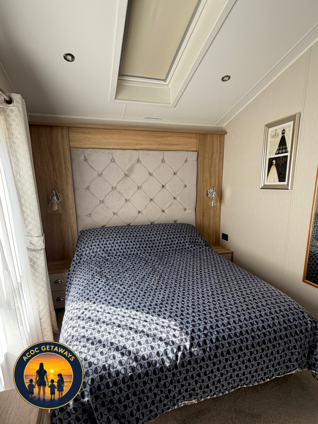 Hotel bedroom with a bed with a decorative headboard, bedside table, wall-mounted lamps, patterned bedding, window with curtain, and framed artwork on the wall.