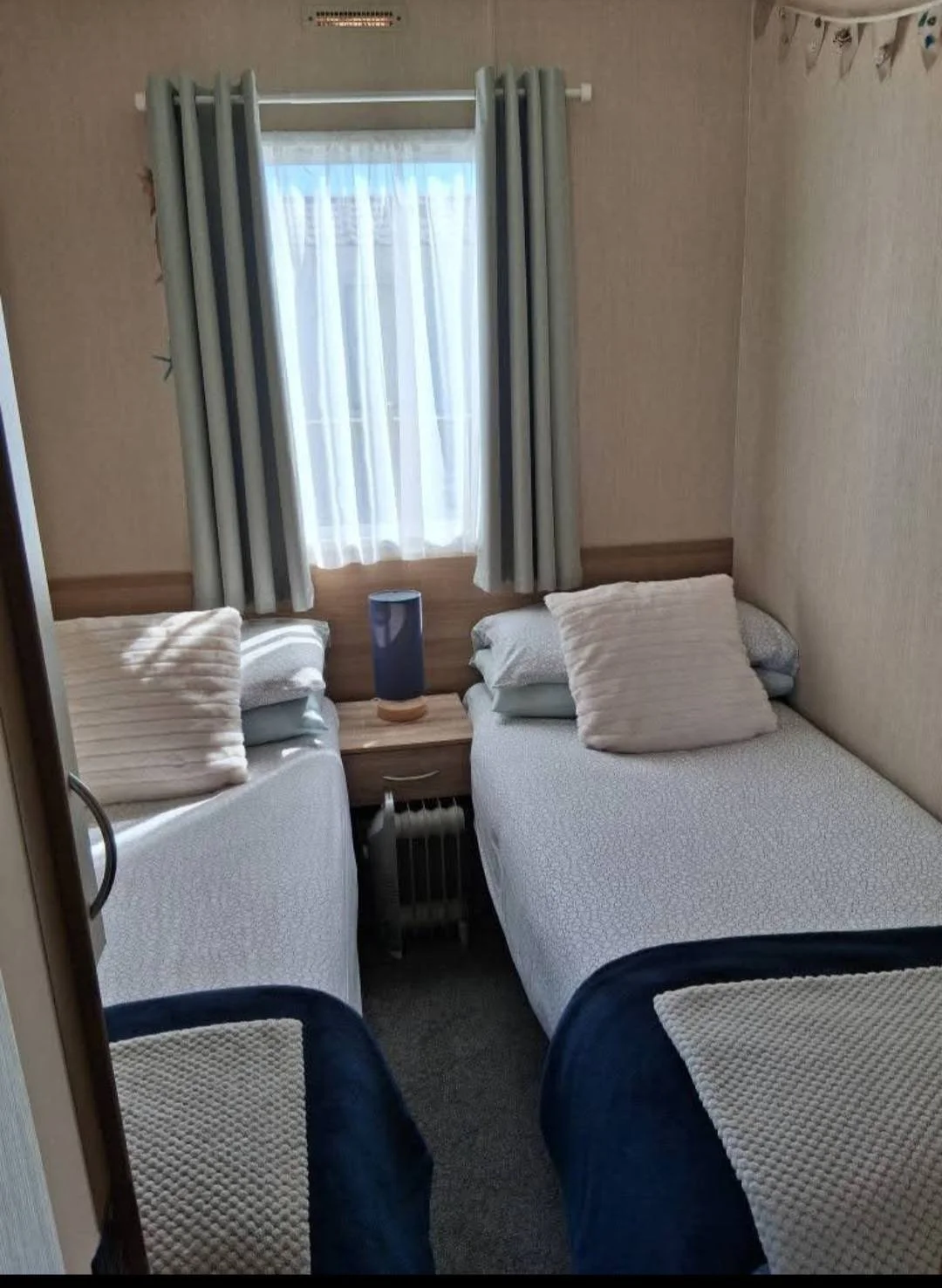 A small bedroom with two twin beds, a window with curtains, a bedside table with a lamp, and two chairs with pillows.