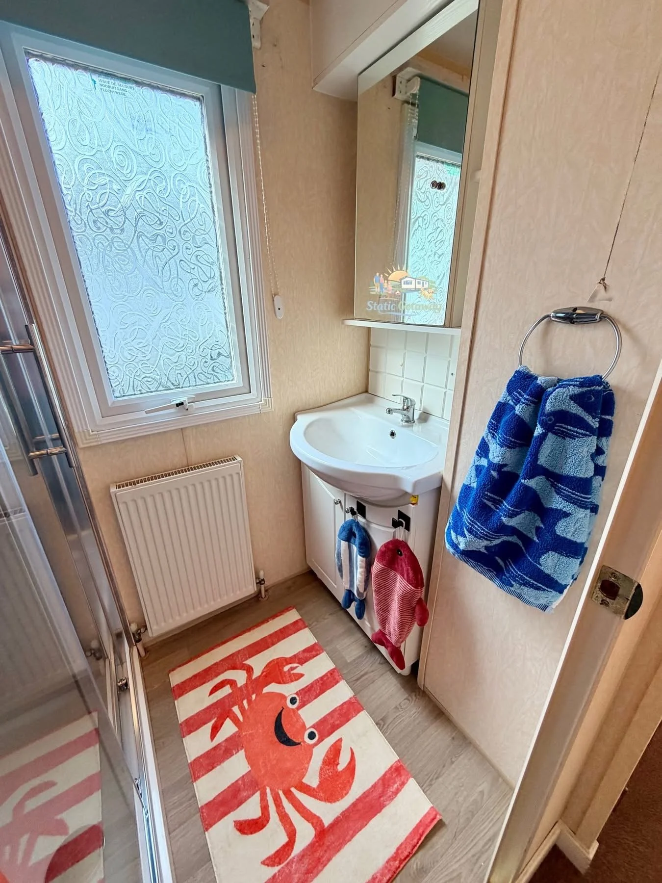 A small bathroom with a window with frosted patterned glass, a radiator below the window, a small white vanity with a sink, a mirror above the sink, and hand towels hanging on a ring and hooks. The floor has a red and white striped rug with a red lob