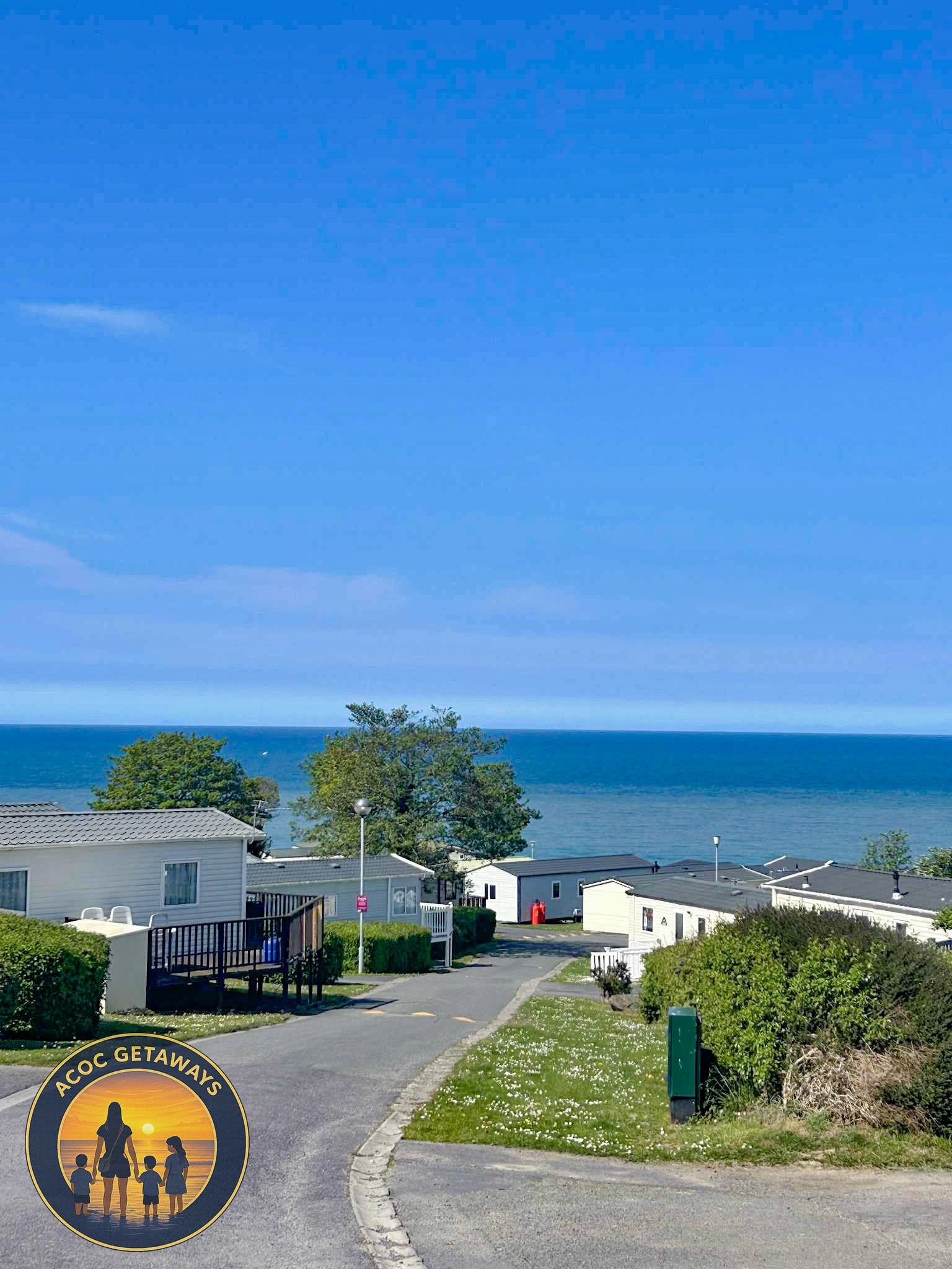 Coastal view of a caravan park with white mobile homes, a winding road, greenery, trees, and the ocean in the background under a bright blue sky.