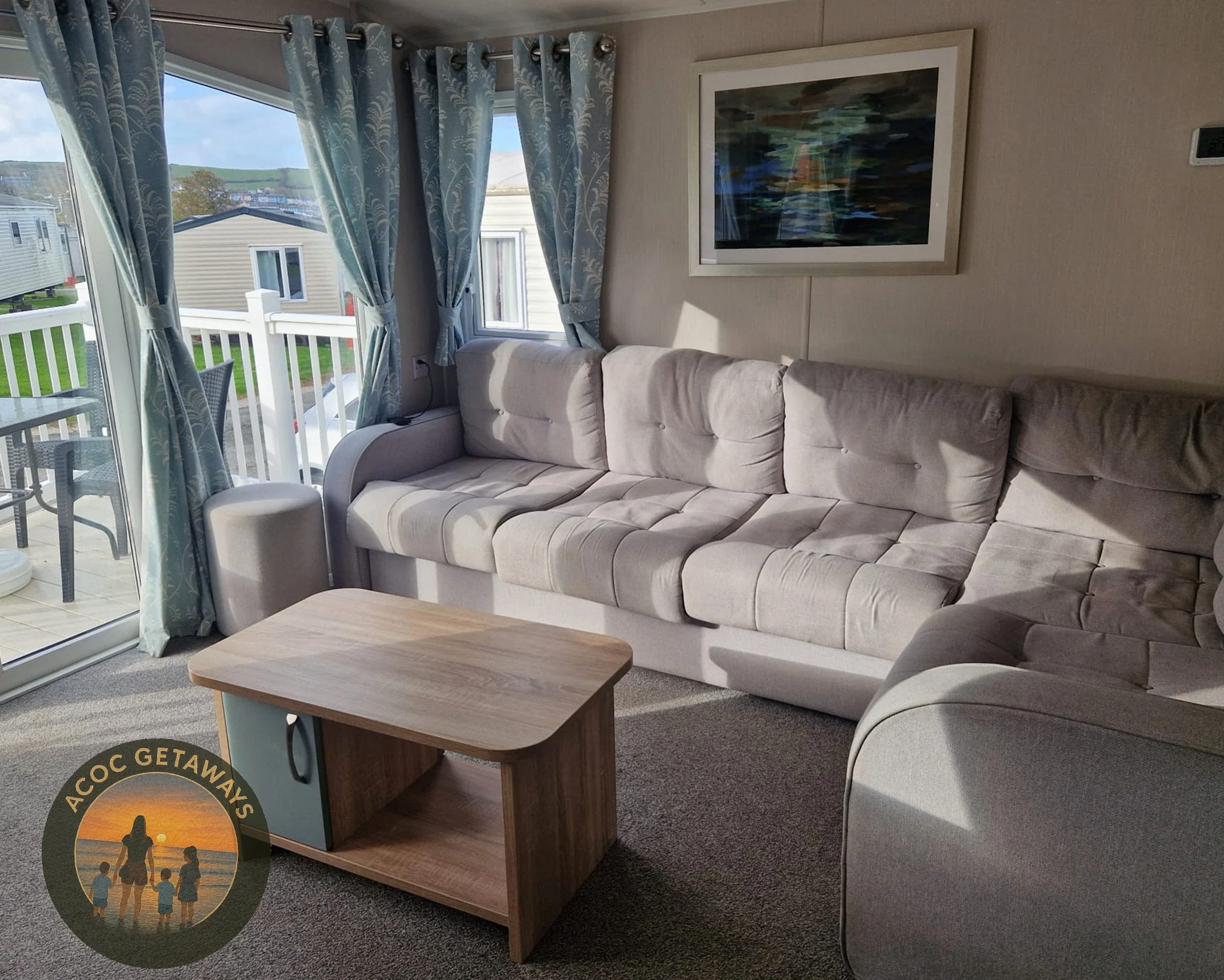 Living room with a large beige couch, a wooden coffee table, a sliding glass door with curtains leading to an outdoor balcony with chairs, a framed abstract painting on the wall, and a view of neighboring mobile homes.