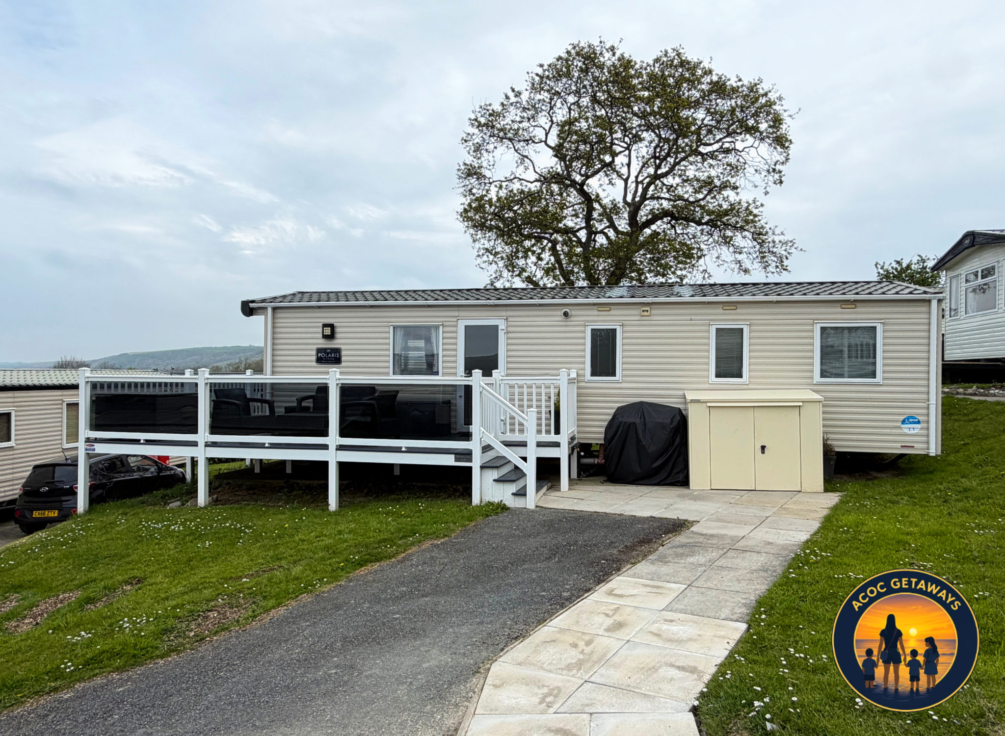 A backyard with a beige mobile home and a wooden deck, a parked black car underneath, a paved walkway, green grass with small white flowers, a large tree behind the house, and cloudy sky.