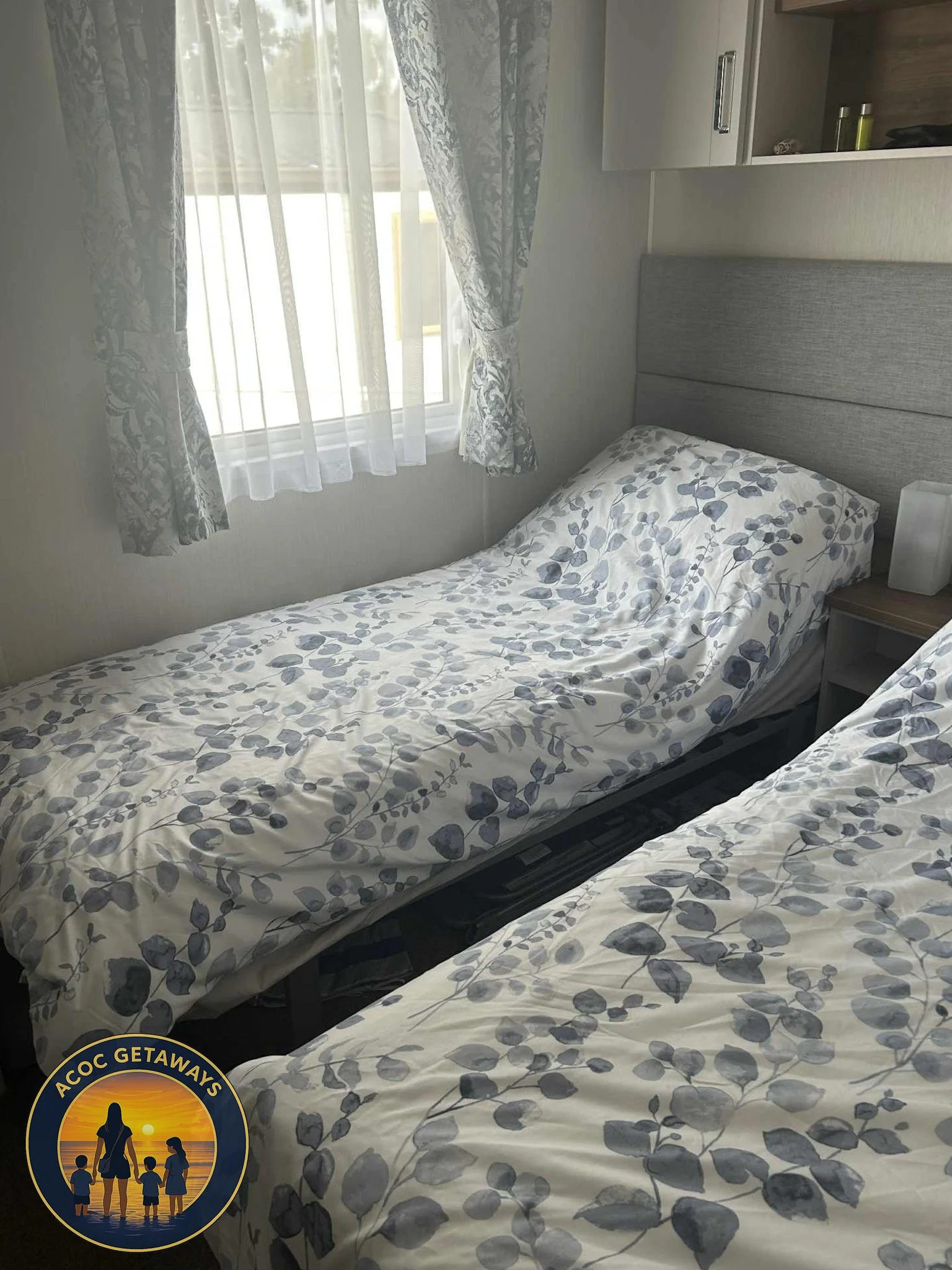 Two single beds with white and gray floral bedspreads in a room with a window and gray headboards.
