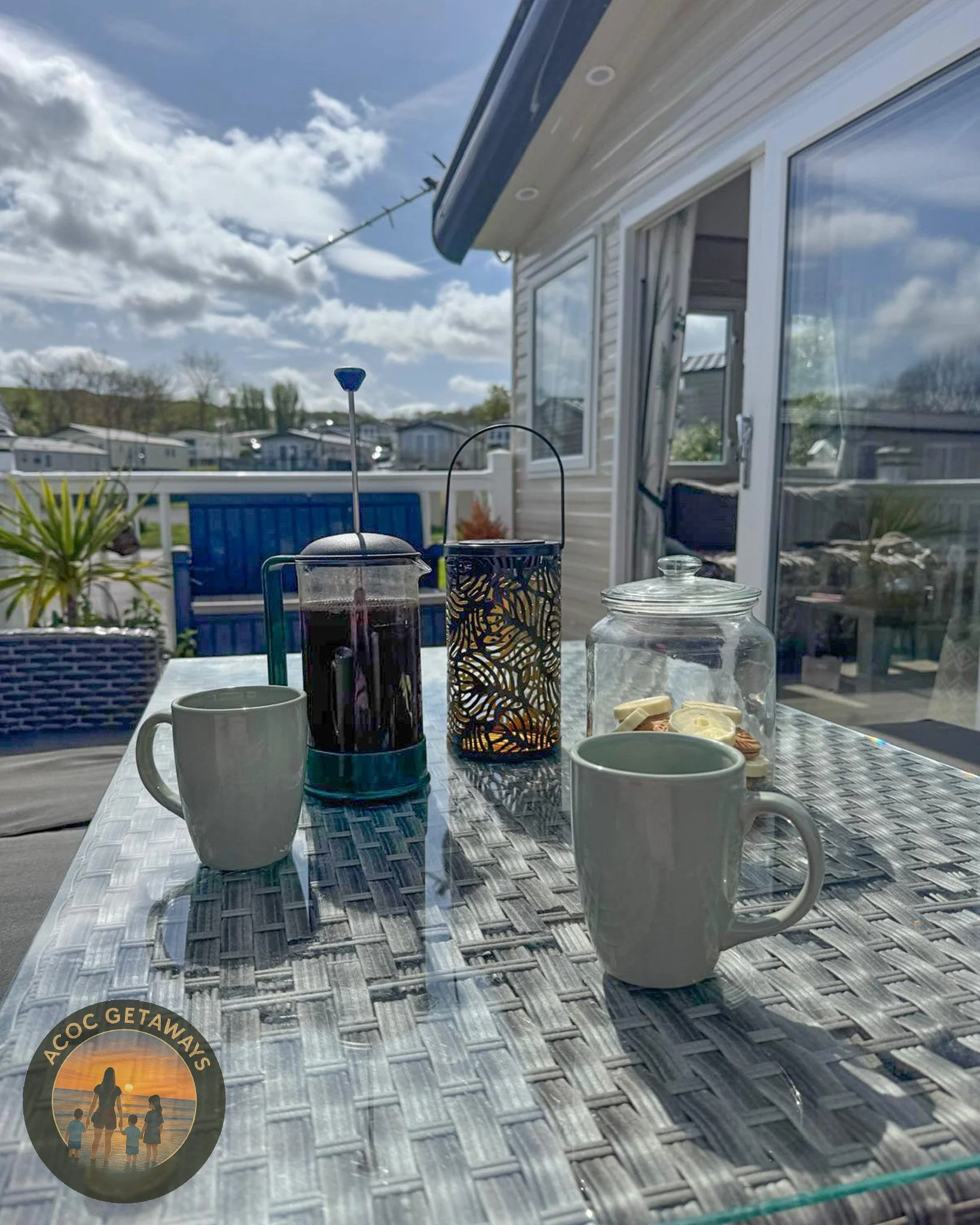 An outdoor patio table with two white coffee mugs, a French press, a decorative candle holder, and a glass jar with snacks, set against a sunny sky and neighboring houses.
