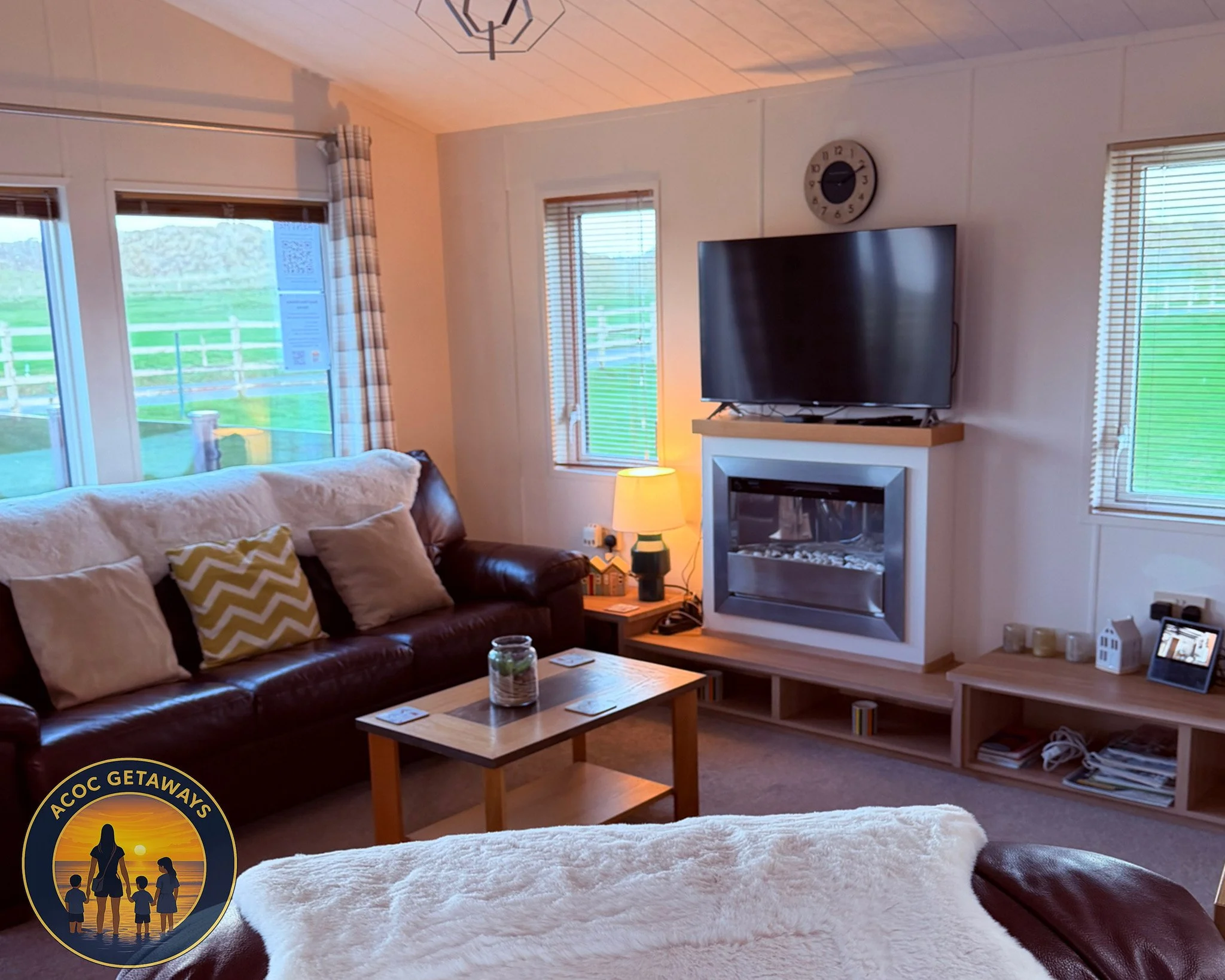 Cozy living room with a brown leather sofa with beige and green chevron pillows, a wooden coffee table with a small plant in a glass jar, a bedside table with a lamp, a wall-mounted TV above a modern fireplace, large windows with blinds and checkered