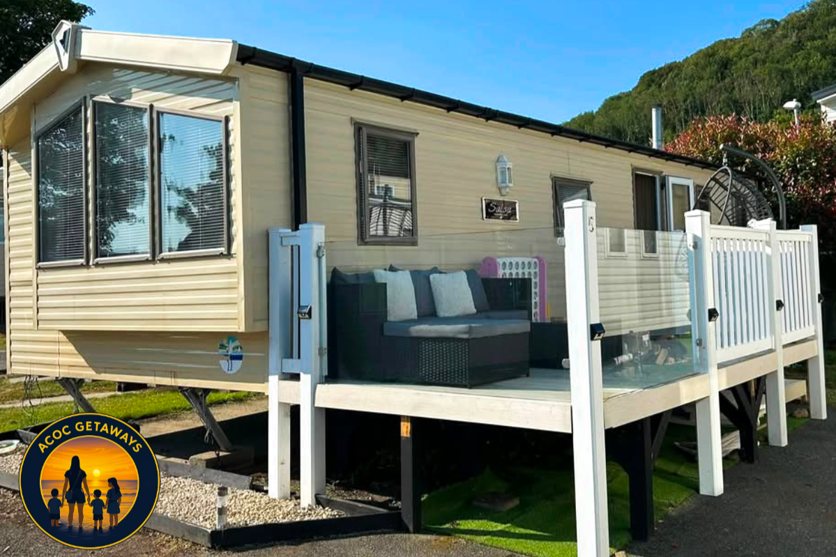 A beige mobile home with a small porch and outdoor seating, surrounded by greenery under a blue sky.