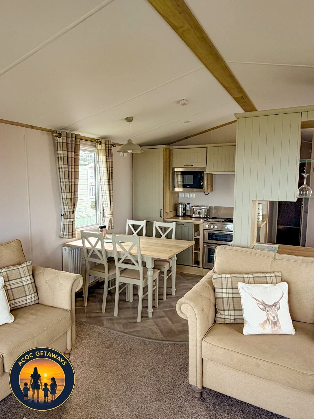 Interior of a cozy living area with beige sofas, plaid cushions, a wooden dining table with six chairs, a window with plaid curtains, and a small kitchen with appliances.